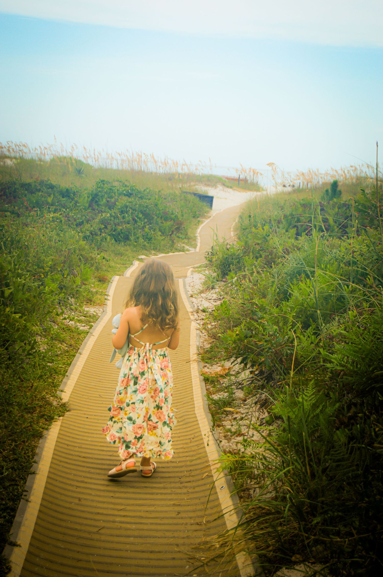 Wooden boardwalk winding through sand dunes with beachgoers in the distance.