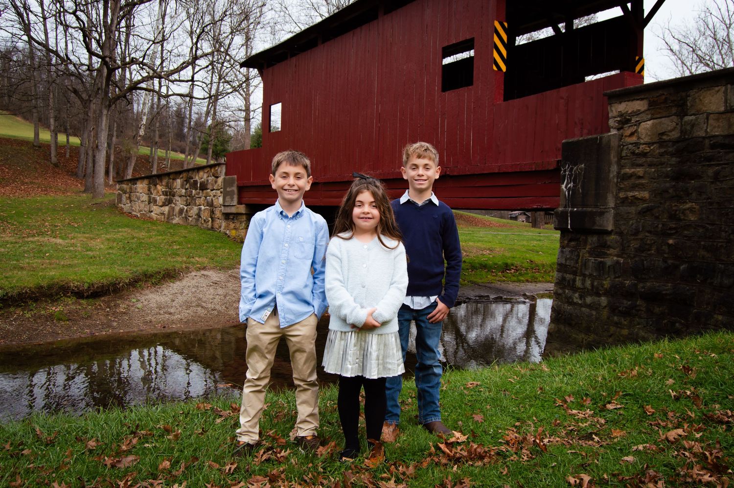 Two children stand in front of a rustic red barn on an autumn day in the countryside.