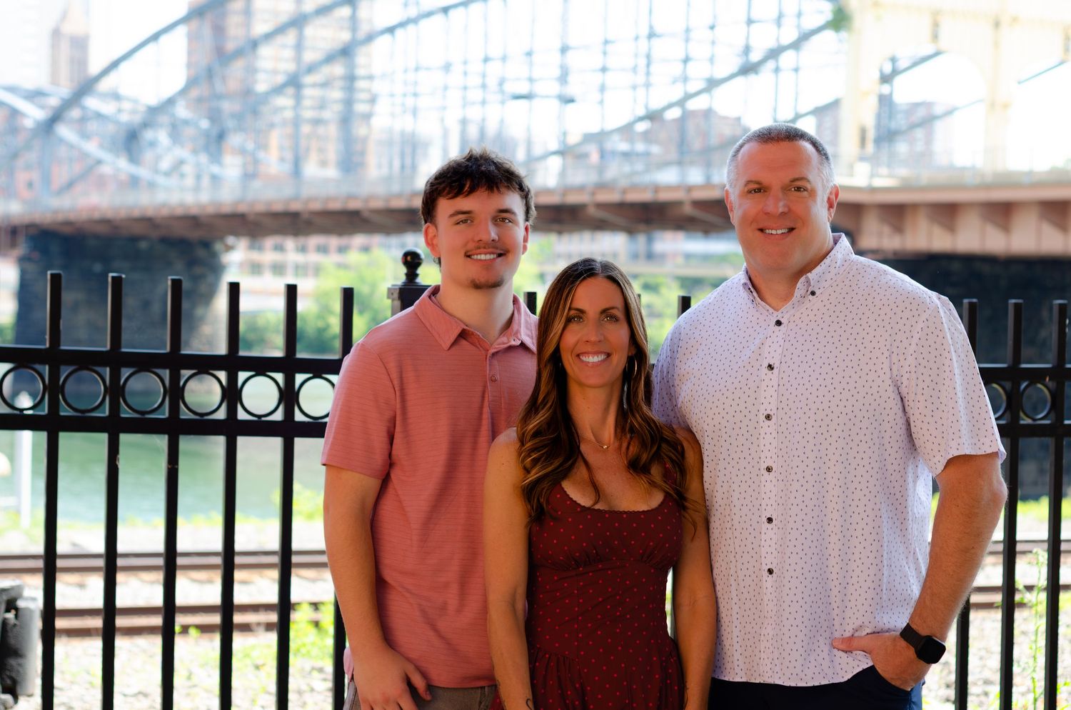 Three people smiling outdoors near a decorative iron fence and bridge, posing for a family portrait on a sunny day.