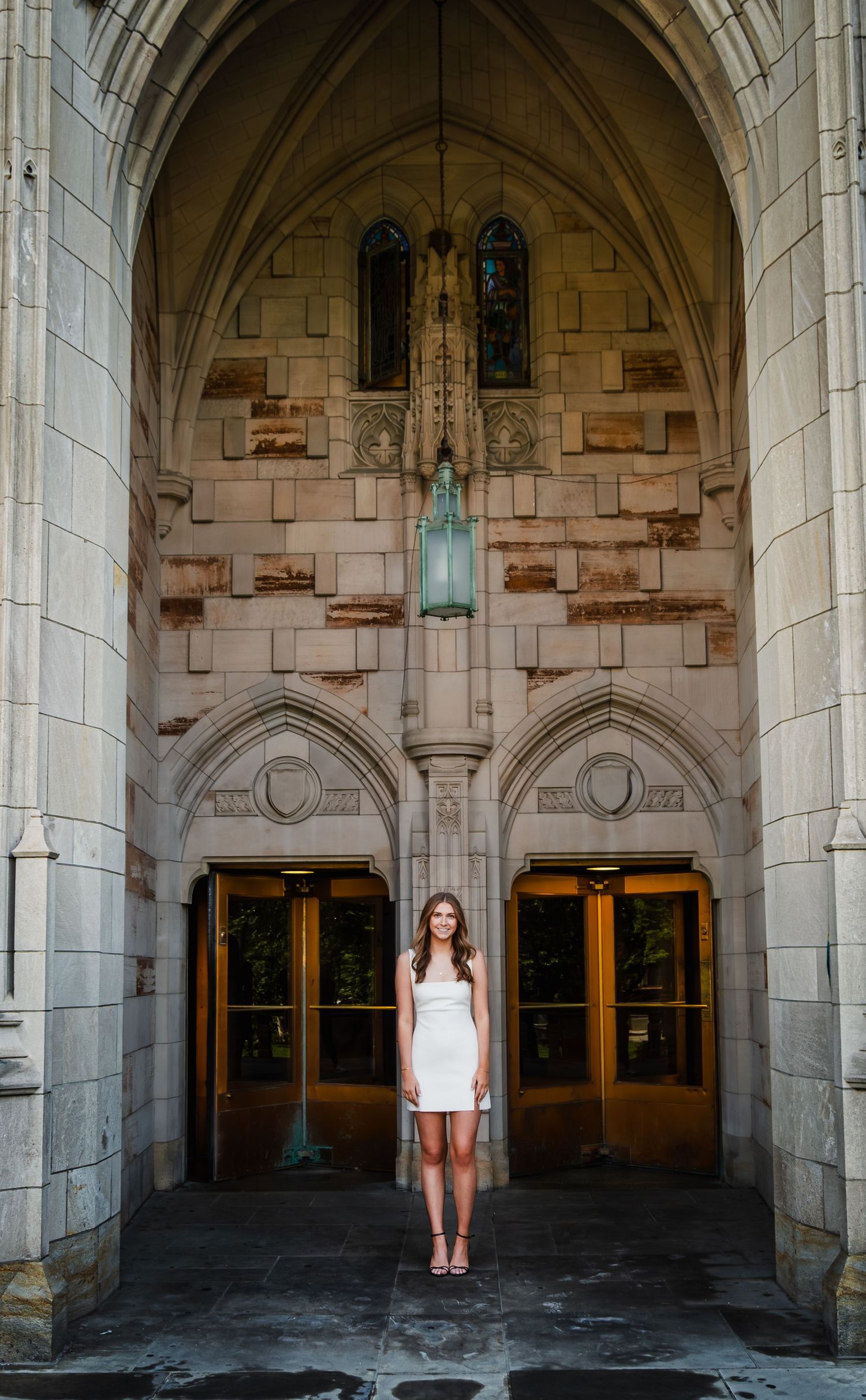 Young woman in white dress standing under Gothic stone archway entrance of historic building.