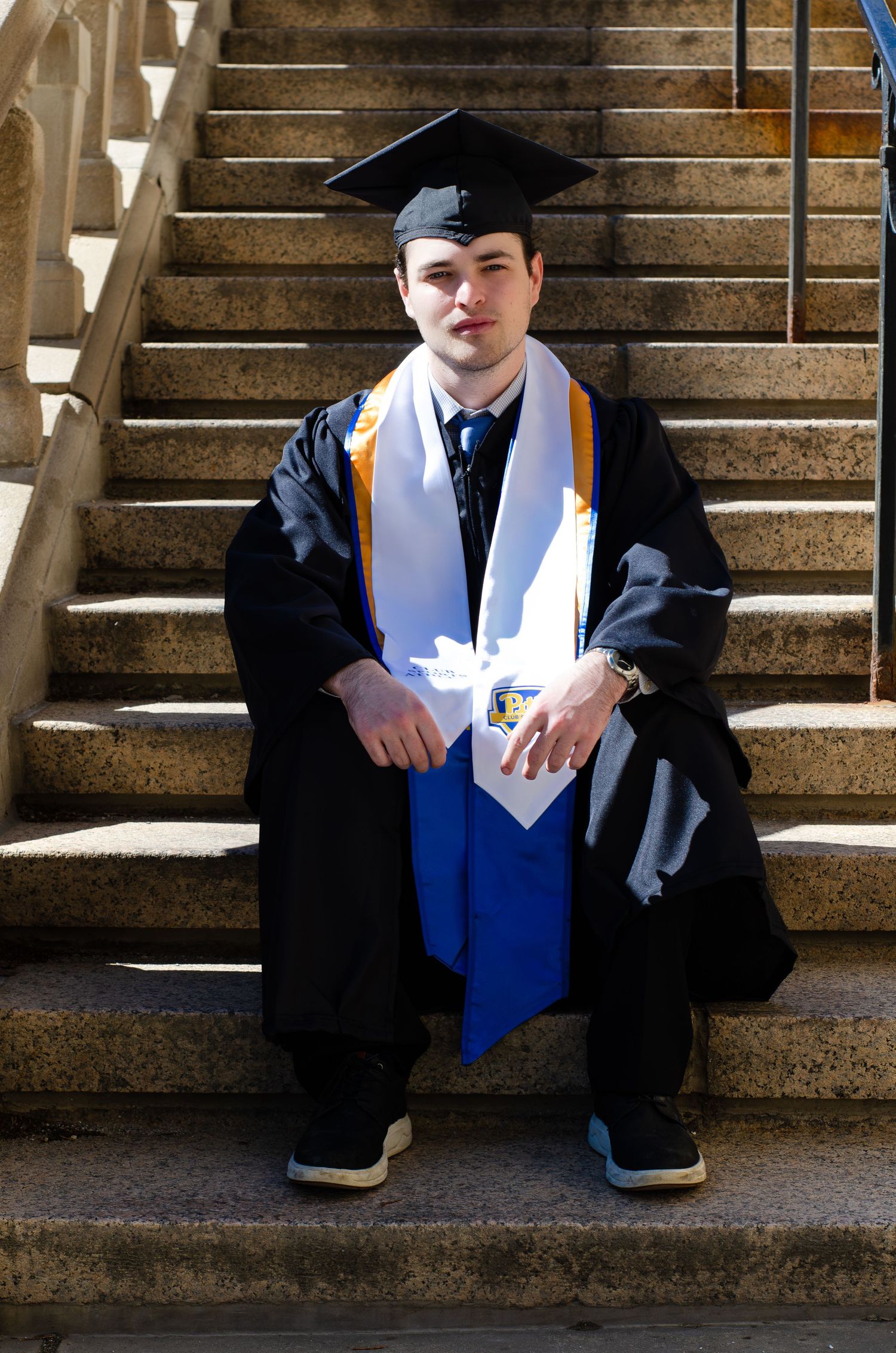 Young man in black graduation gown and cap with blue and white stoles sits on stone steps.