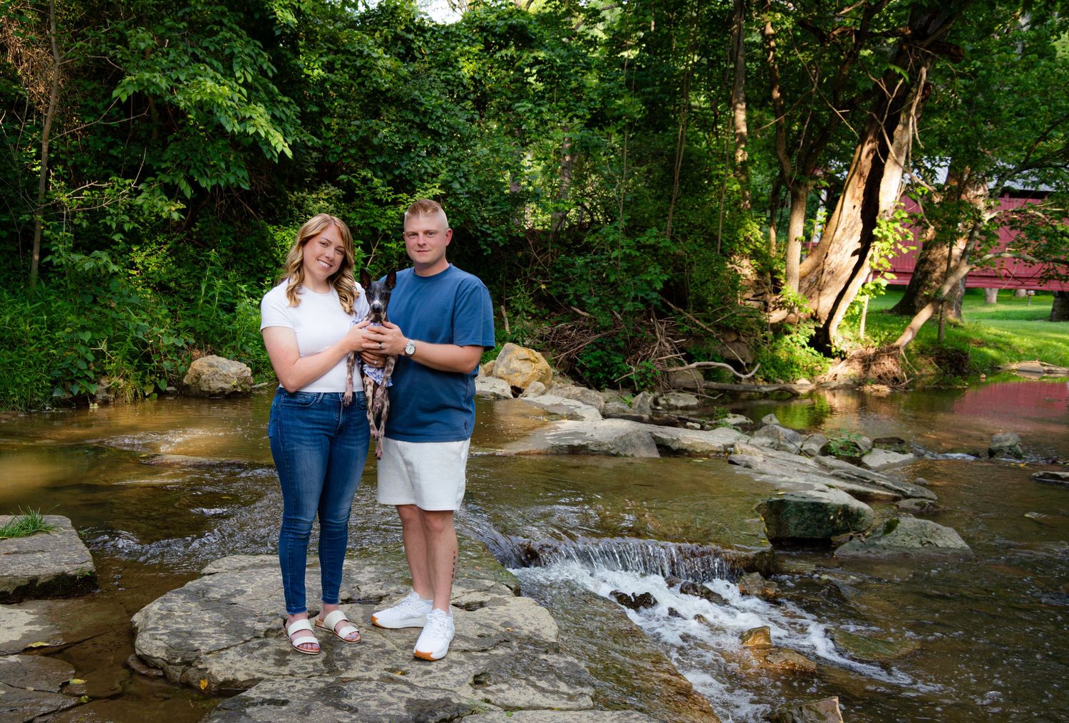 Couple standing on rocks by a scenic creek surrounded by lush green trees in a beautiful outdoor setting.