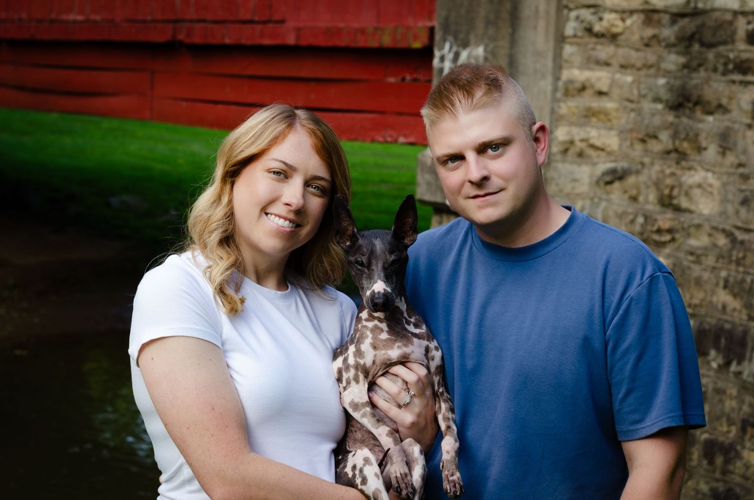 A couple smiles while holding a dog outdoors near a brick wall and red structure.