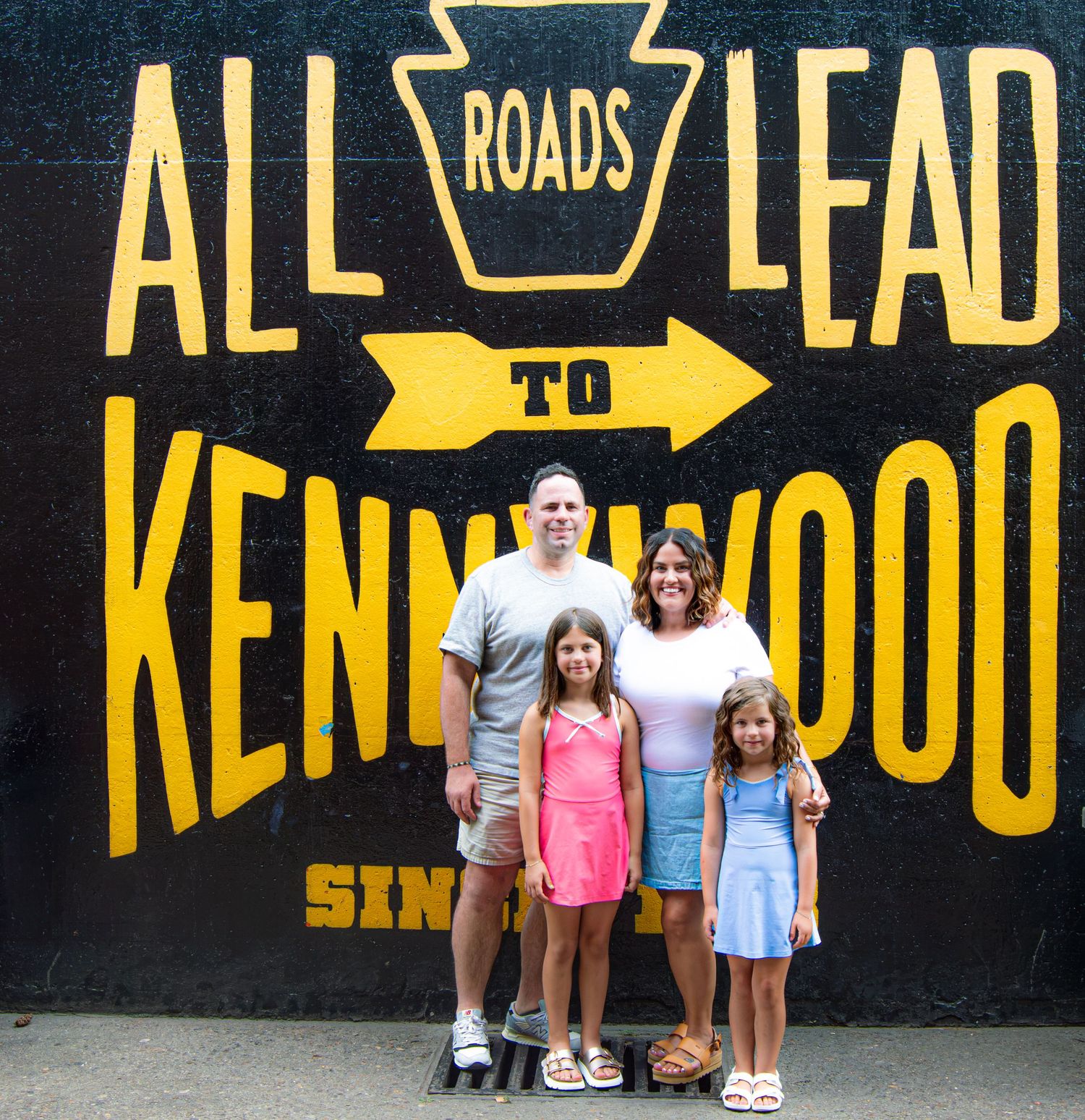 Family of four posing in front of Kennywood amusement park mural reading All Roads Lead to Kennywood.