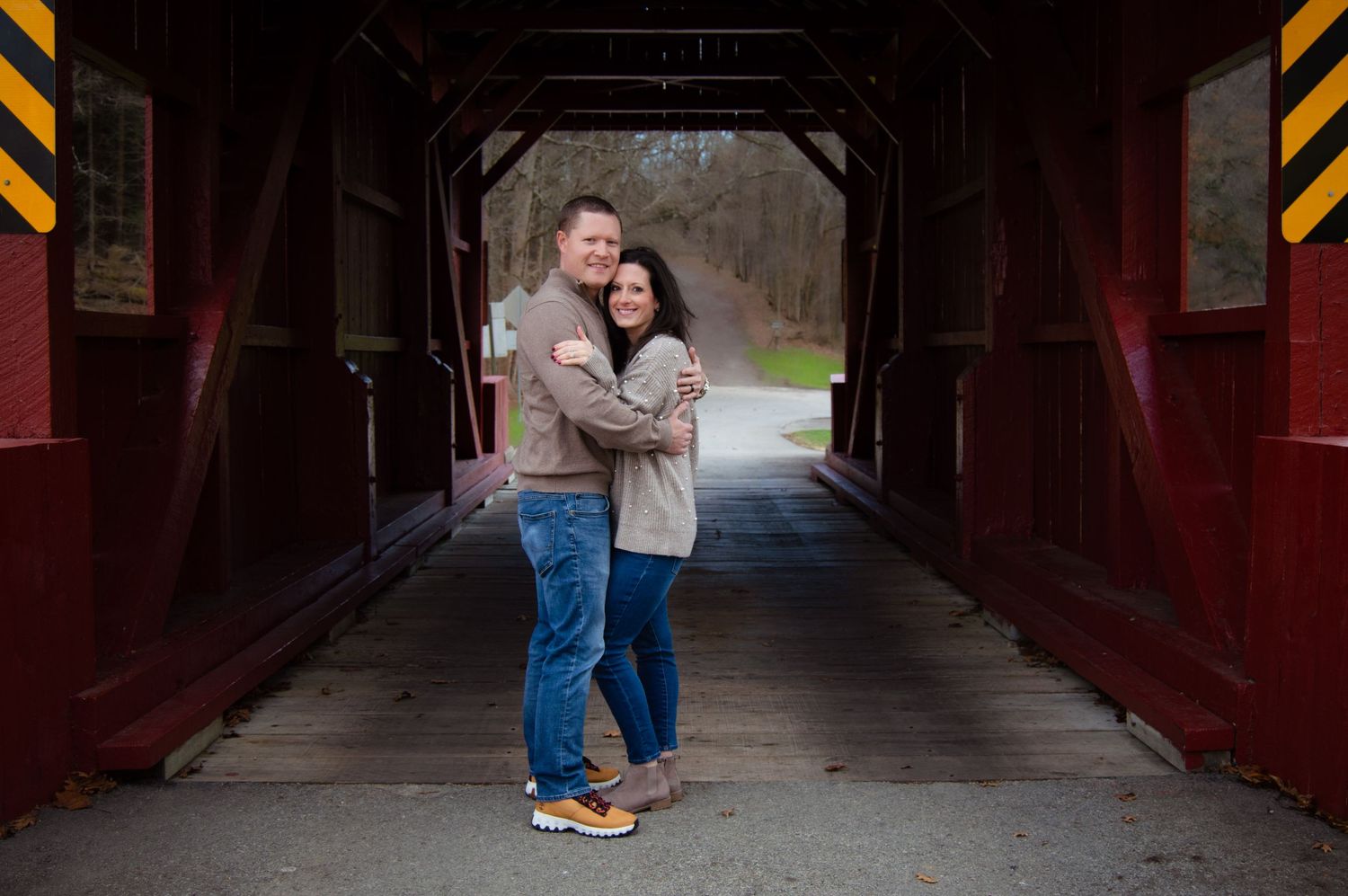 A couple stands together in a covered red bridge walkway during an engagement photo session.