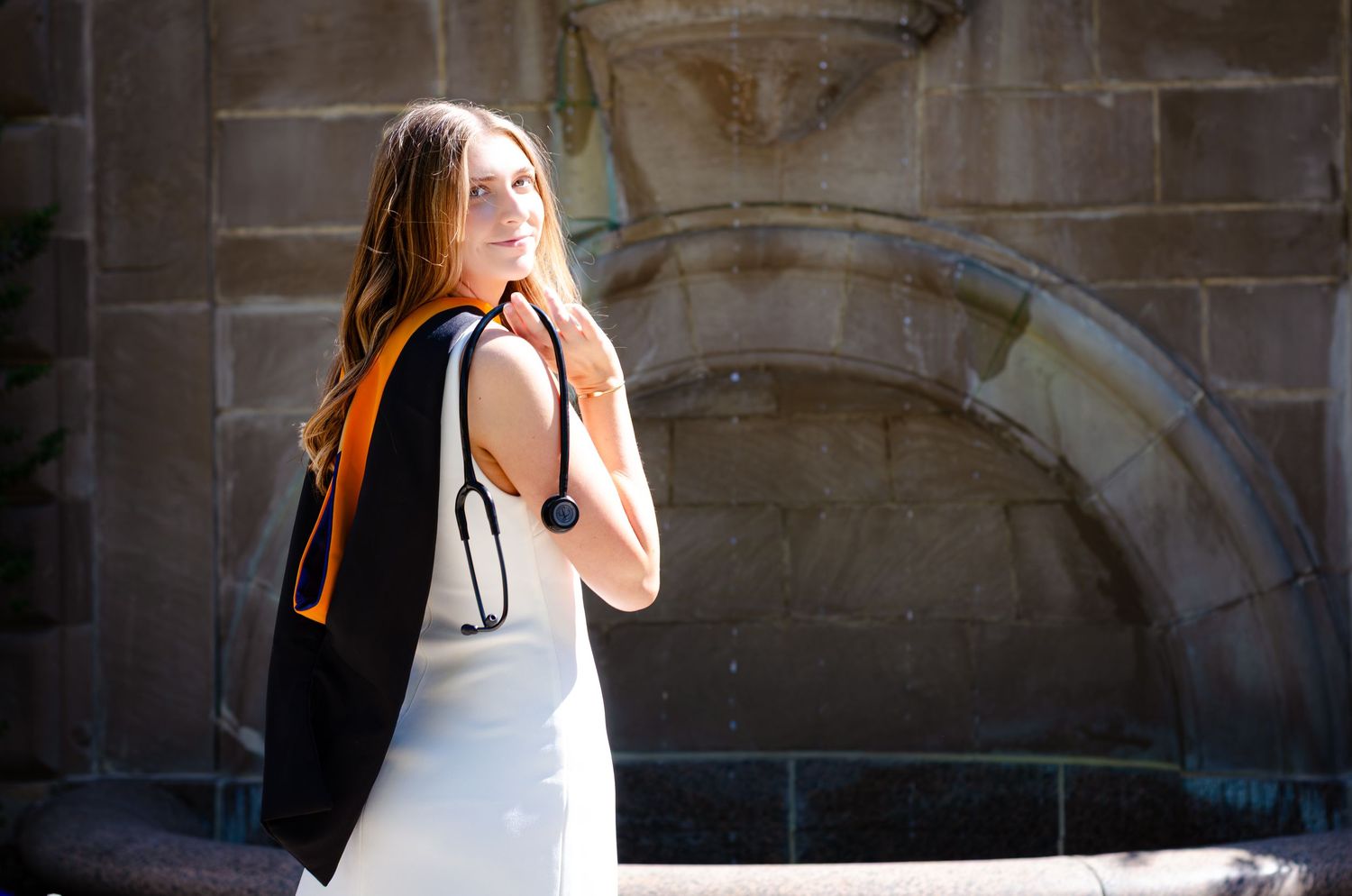 Young woman in white dress with black graduation gown and stethoscope poses by stone fountain.