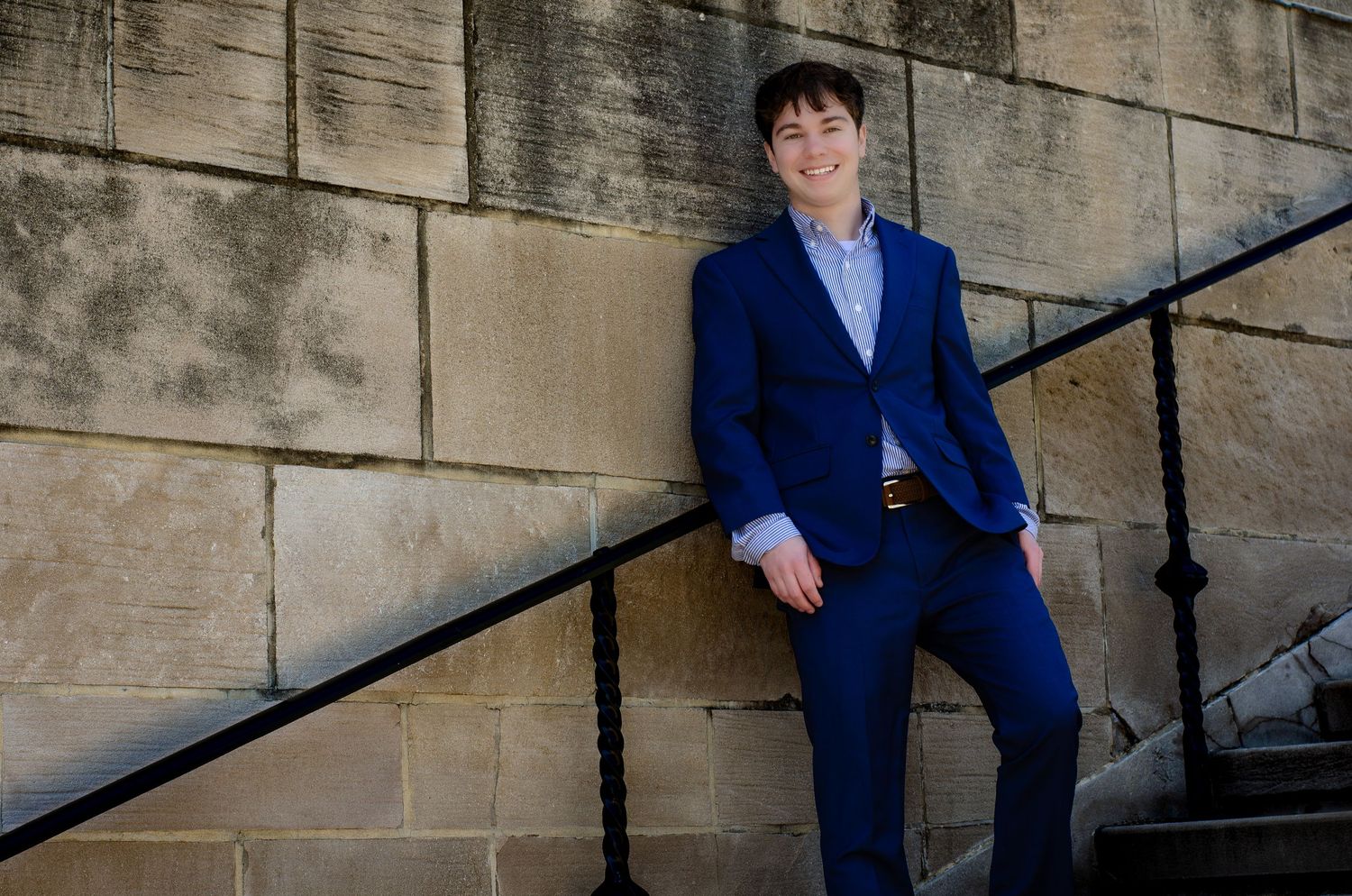Young man in blue suit smiling against stone wall on steps.
