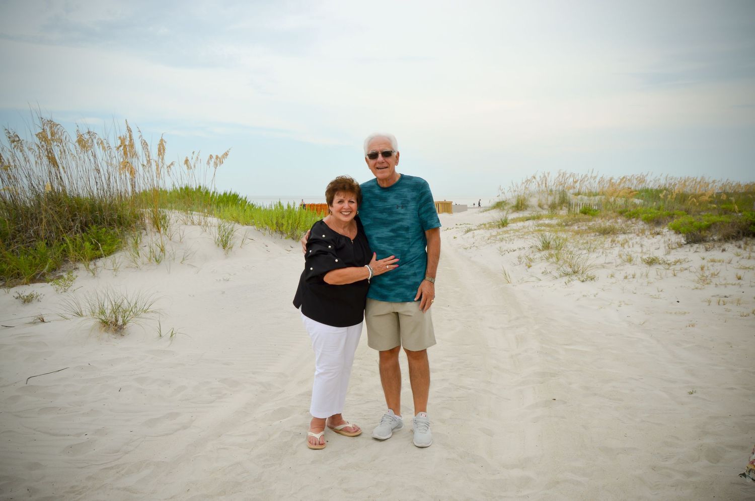 A couple poses together on a sandy beach path surrounded by coastal vegetation.