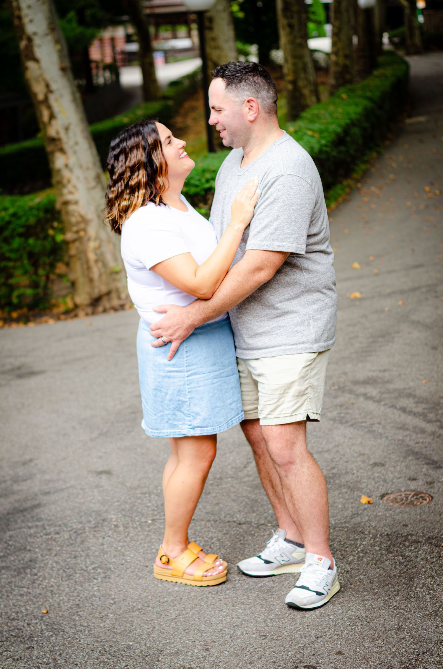 A couple embraces on a tree-lined path, the woman in a white top and blue skirt, the man in a gray shirt and khaki shorts.