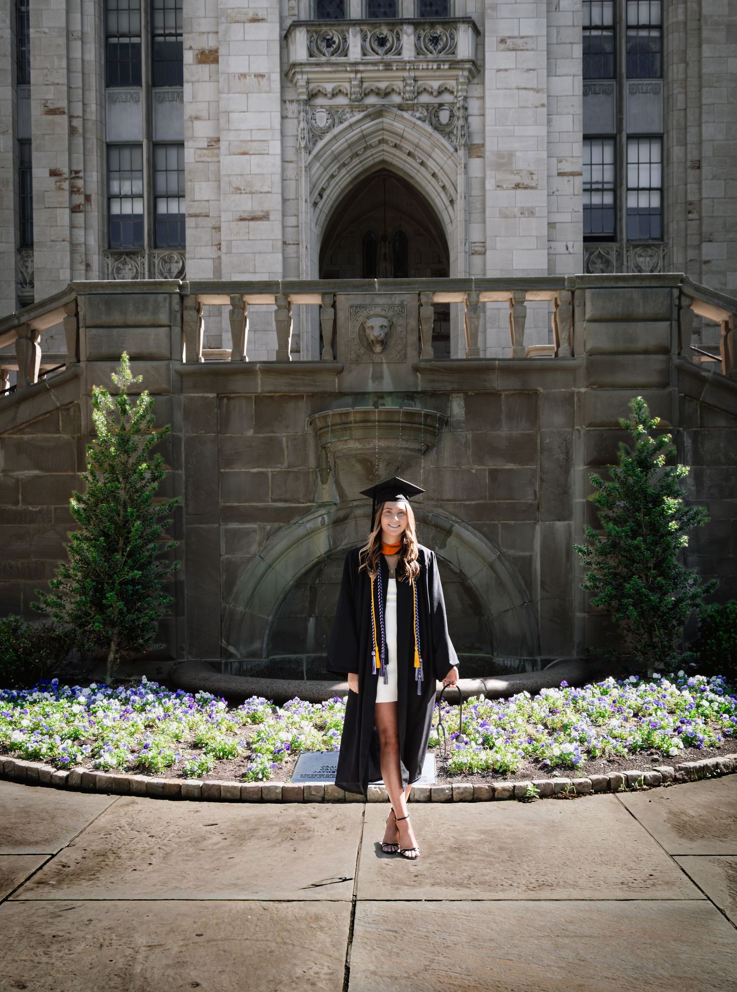 Graduate in cap and gown standing before ornate university building with flower garden.