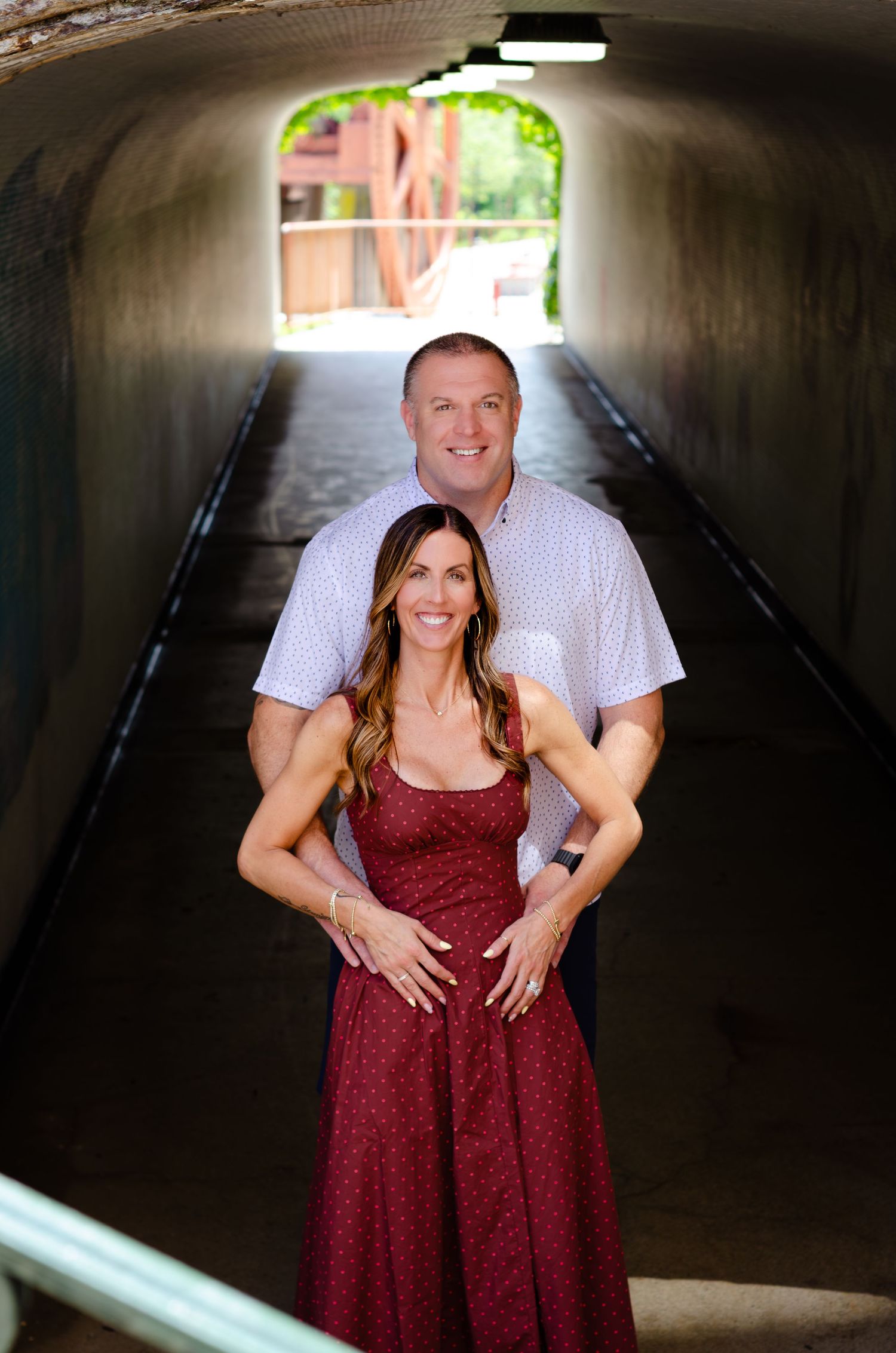 Couple posing in a tunnel, woman in red dress, man in white shirt, bright archway behind them.