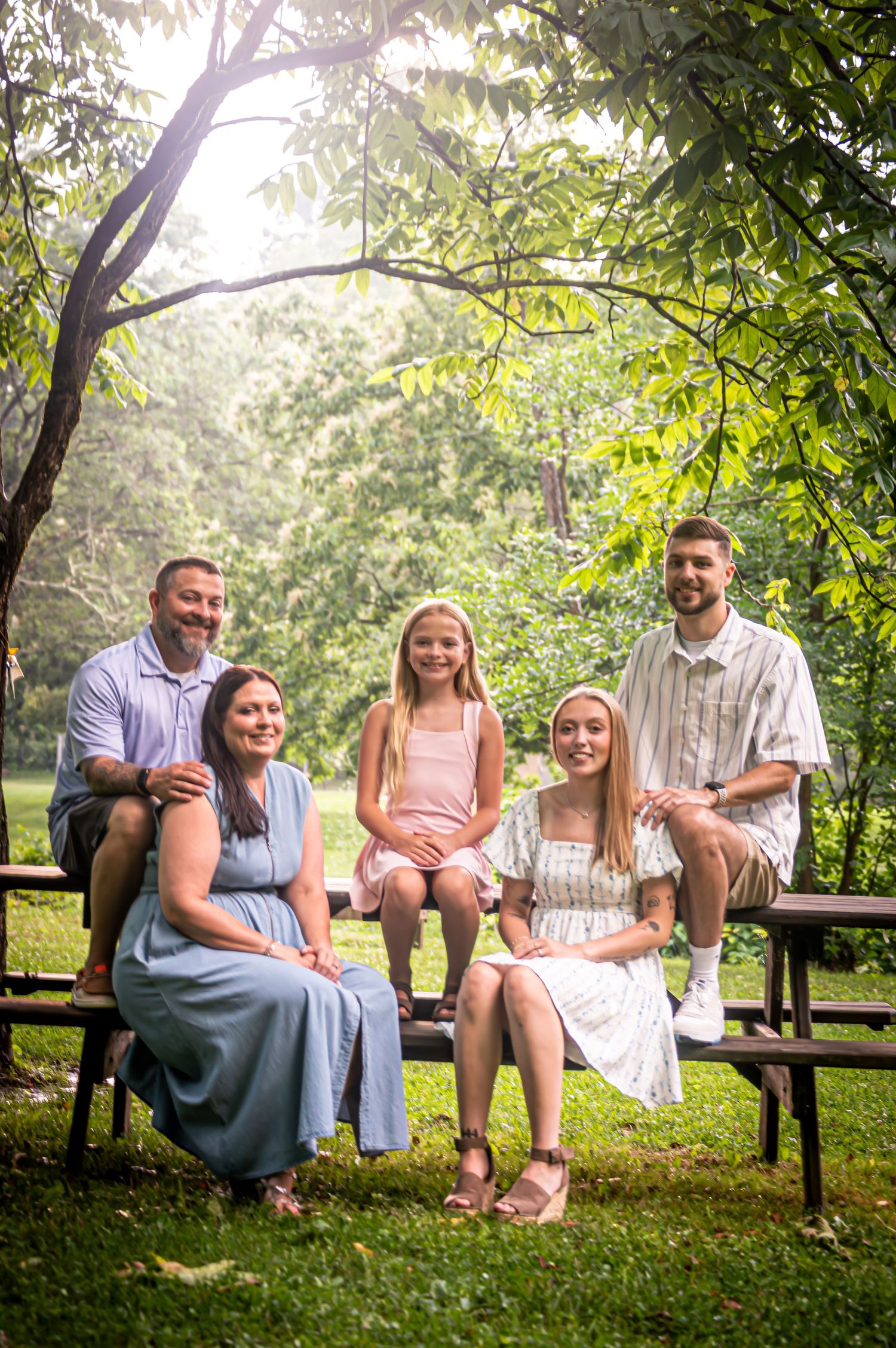 Family group sitting on wooden benches under sunlit trees in a park setting.