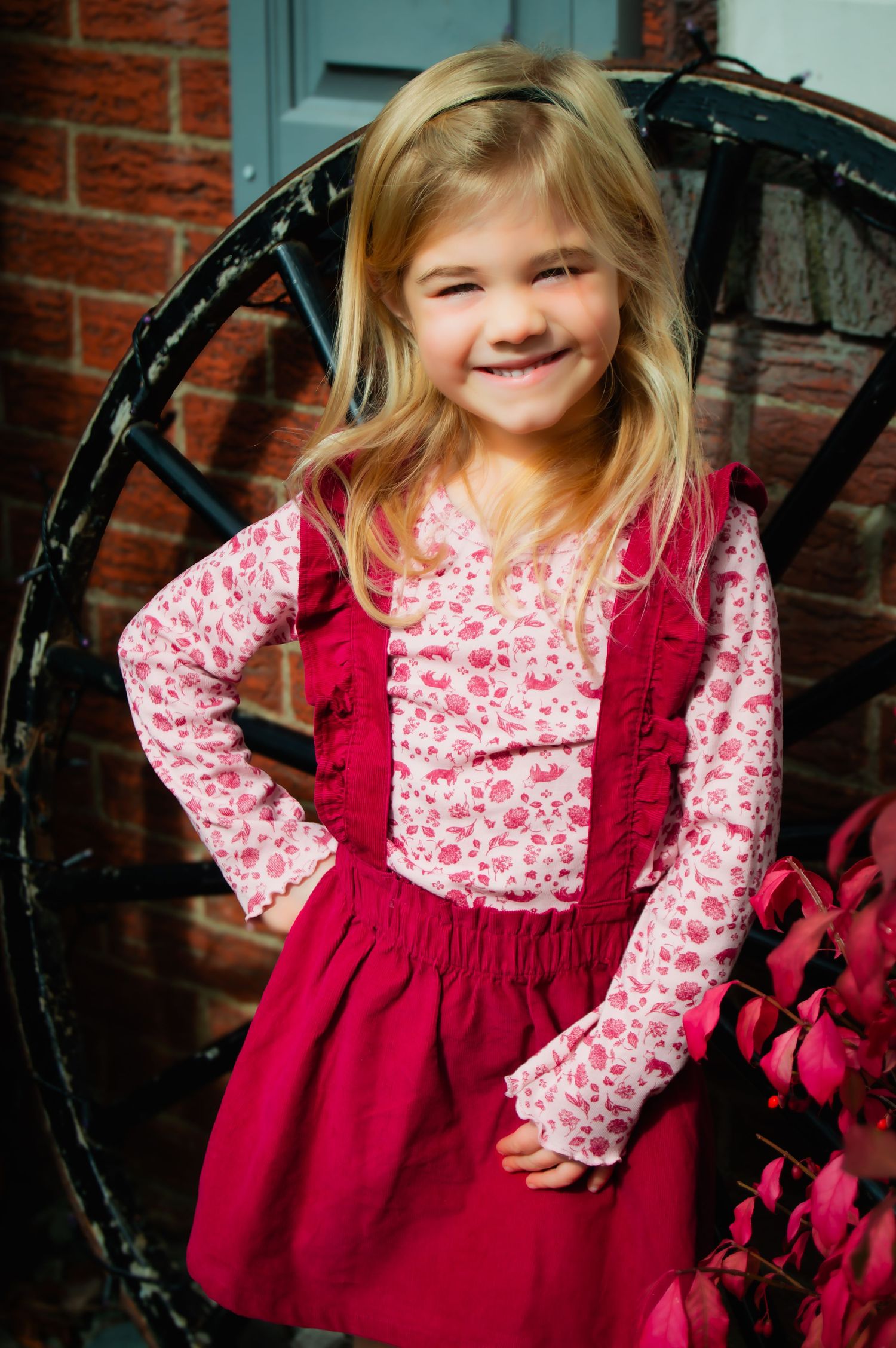 Child wearing red pinafore dress with floral print long sleeves poses against brick wall and black wagon wheel.