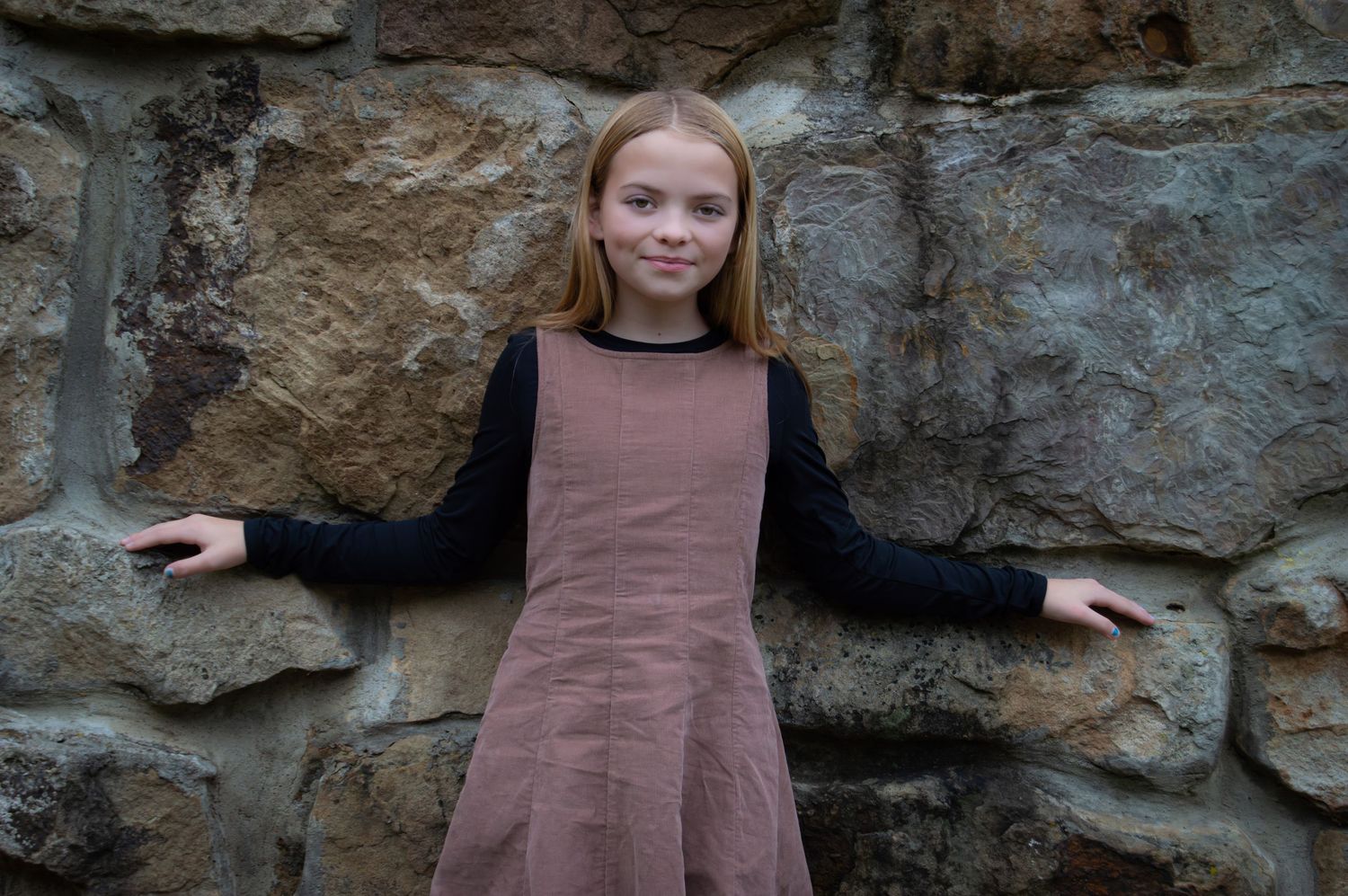 A person wearing a brown dress stands in front of a rocky stone wall outdoors.