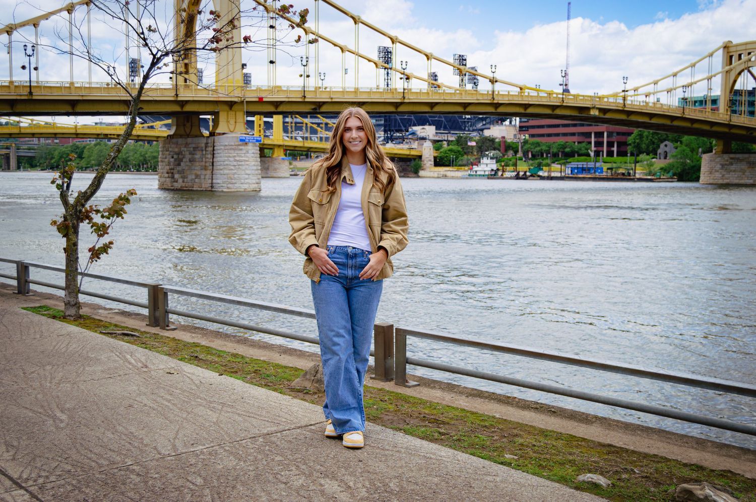 Young woman in tan jacket and flared jeans posing along riverfront with yellow suspension bridge behind her.