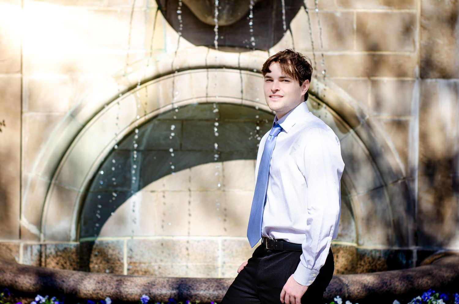 Young man in white dress shirt and blue tie posing confidently in front of an ornate stone fountain.