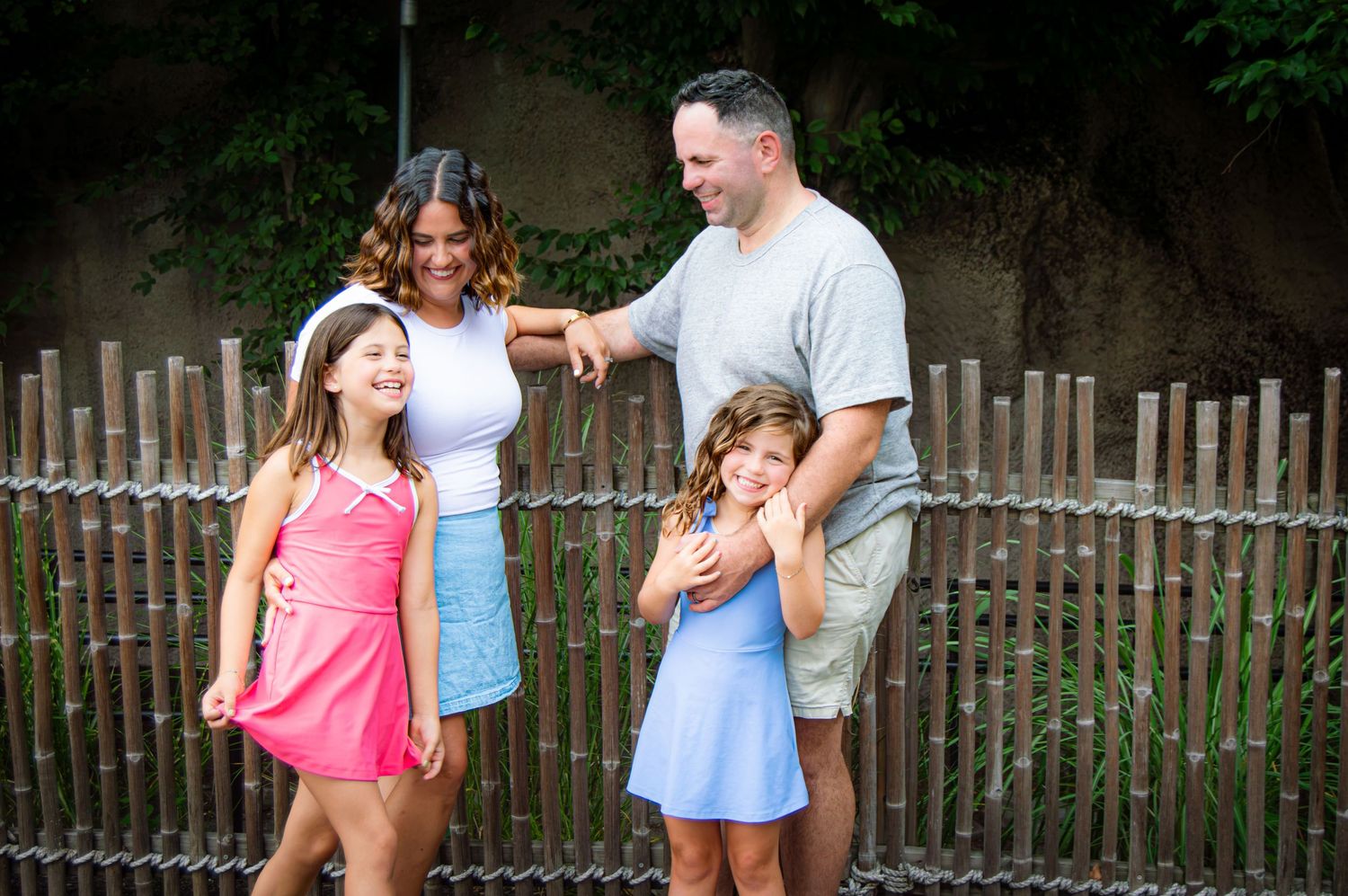 Happy family of four smiling outdoors near a wooden fence on a sunny day.