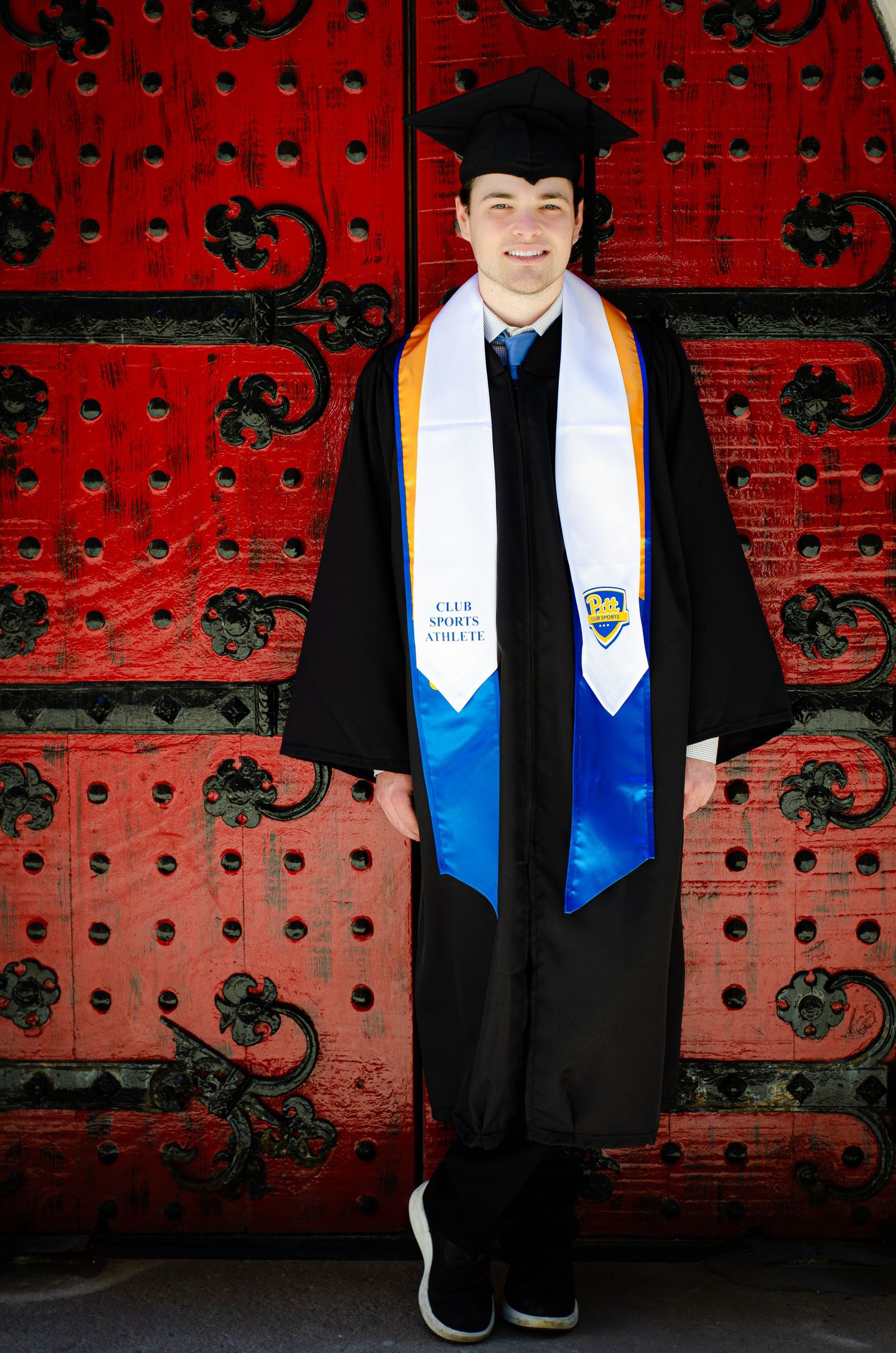 Smiling graduate in cap and gown with blue and white stoles before red doors.