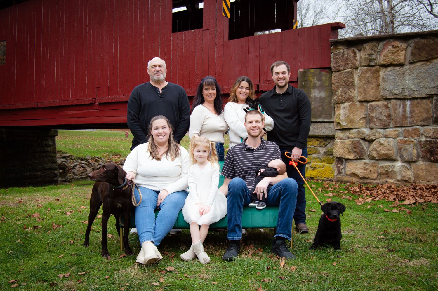 A group of people and dogs sit together for outdoor family portraits against a rustic stone wall and red barn.