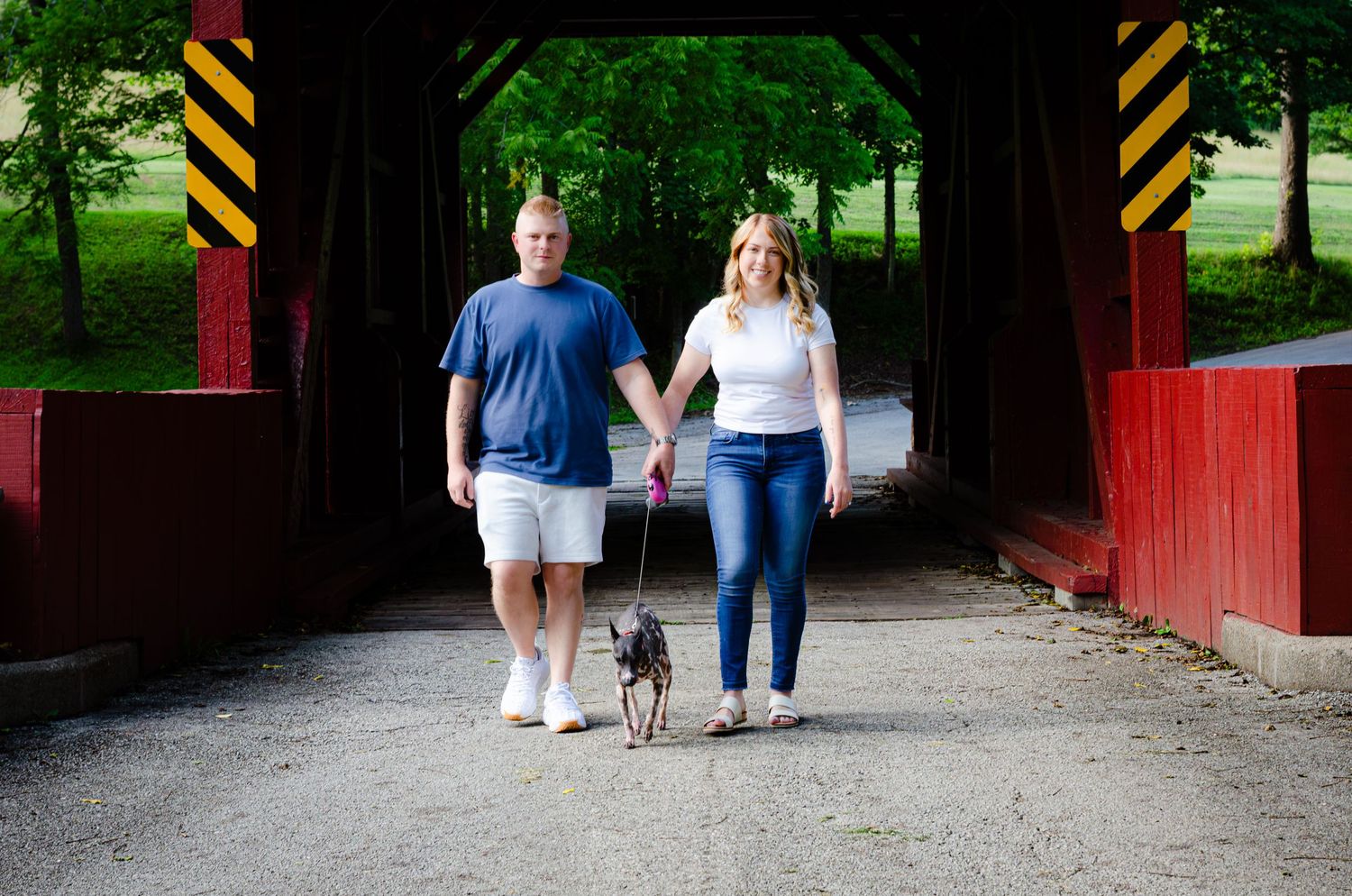 Couple holding hands walking dog through red covered bridge surrounded by lush green trees.