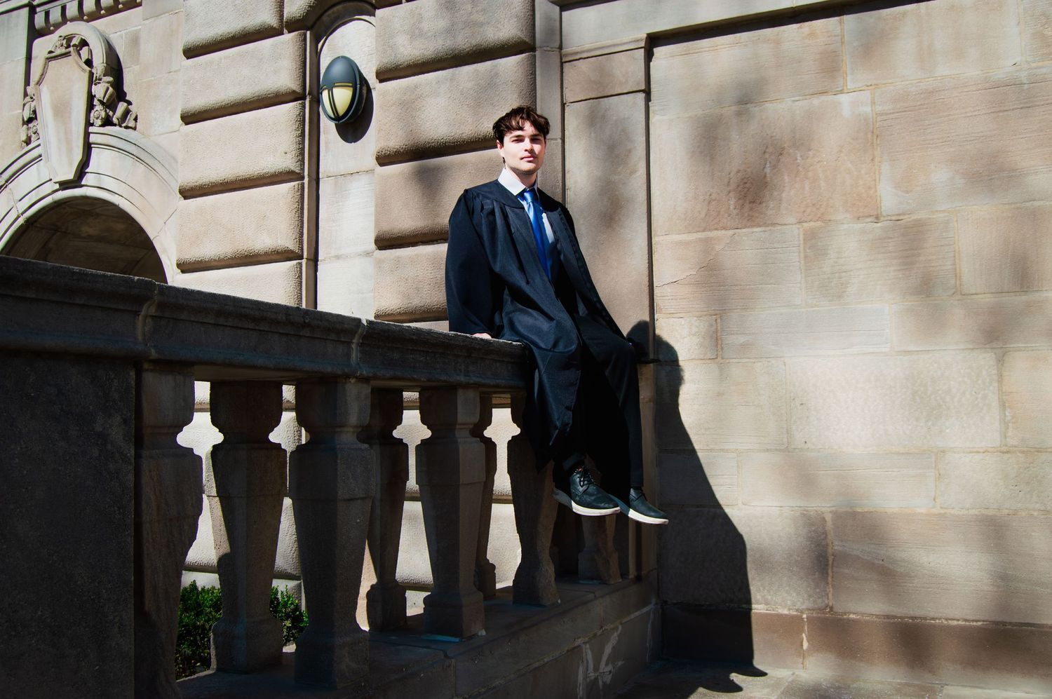 Graduate in black academic gown sitting on stone balustrade of classical building.