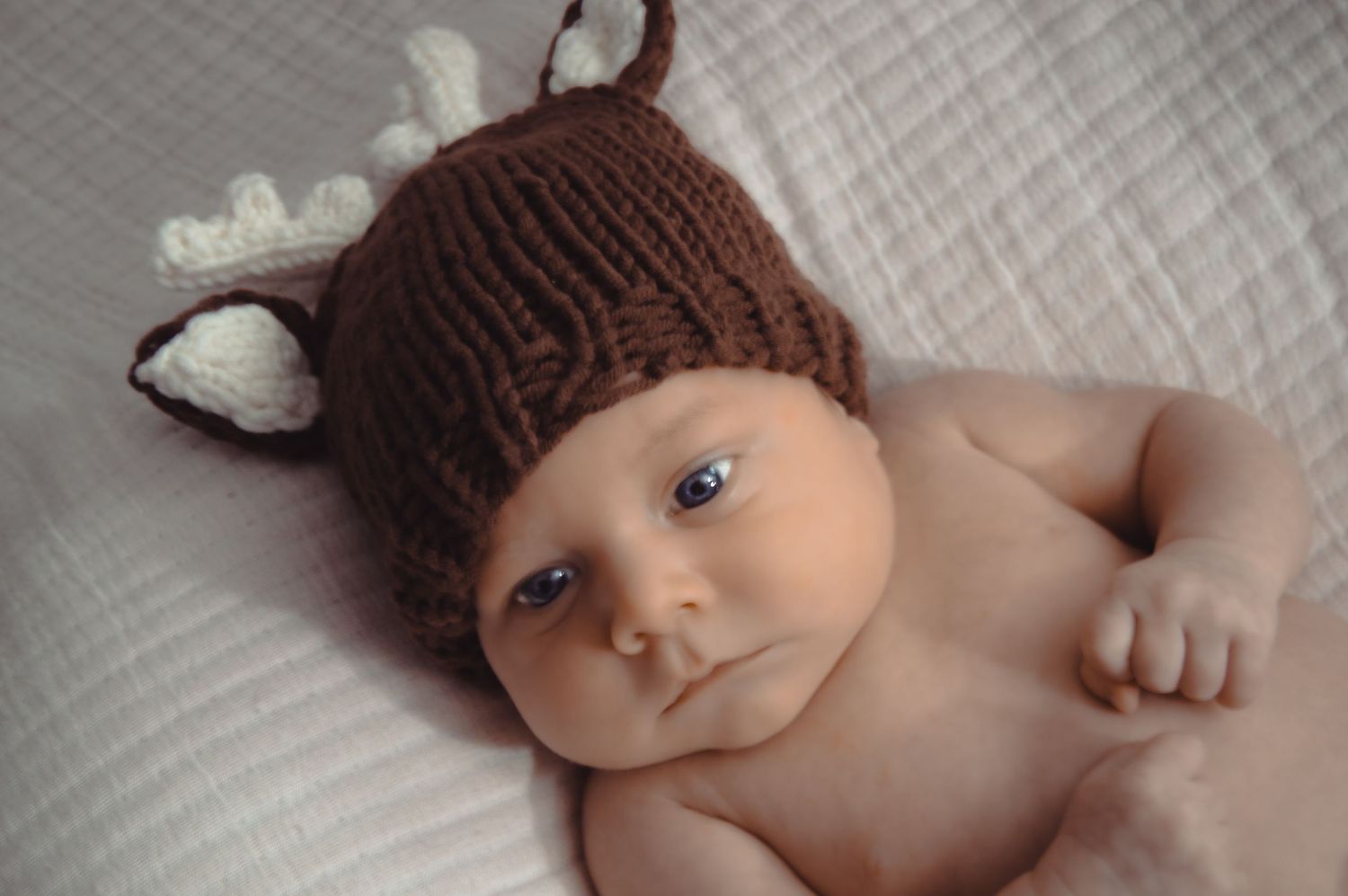 A newborn baby wearing a brown knit hat lays on white fabric for a professional portrait session.