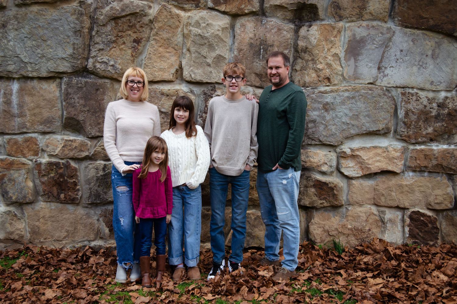 A group stands against a rustic stone wall backdrop during an autumn day with fallen leaves on the ground.