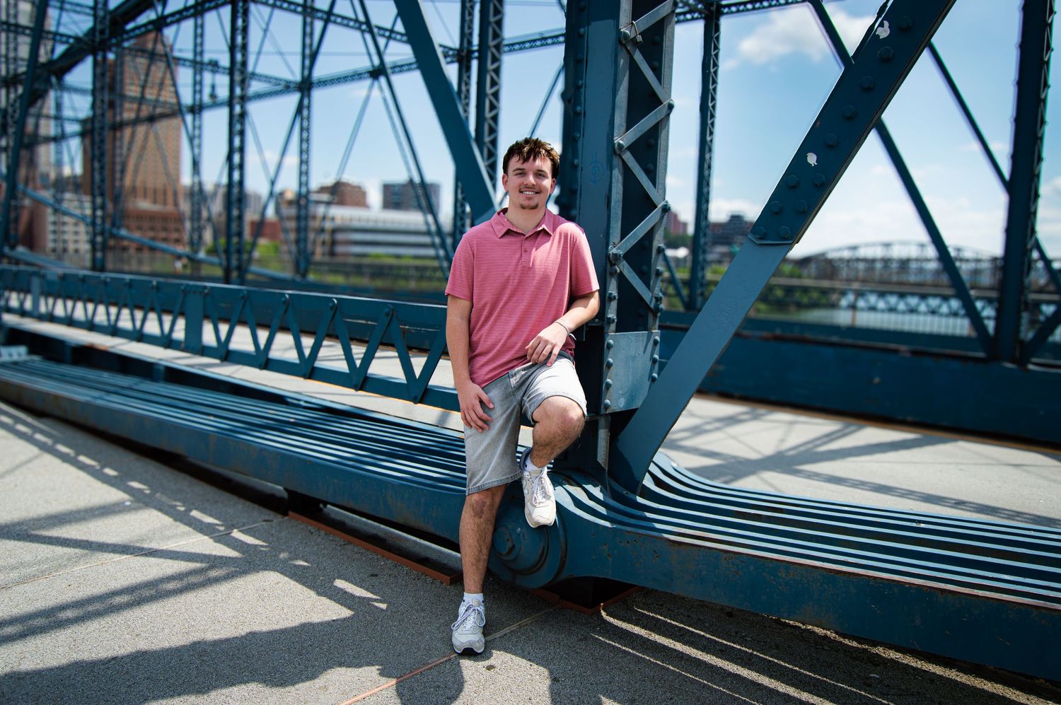 Young man in pink polo and shorts leaning against a steel bridge structure on a sunny day.