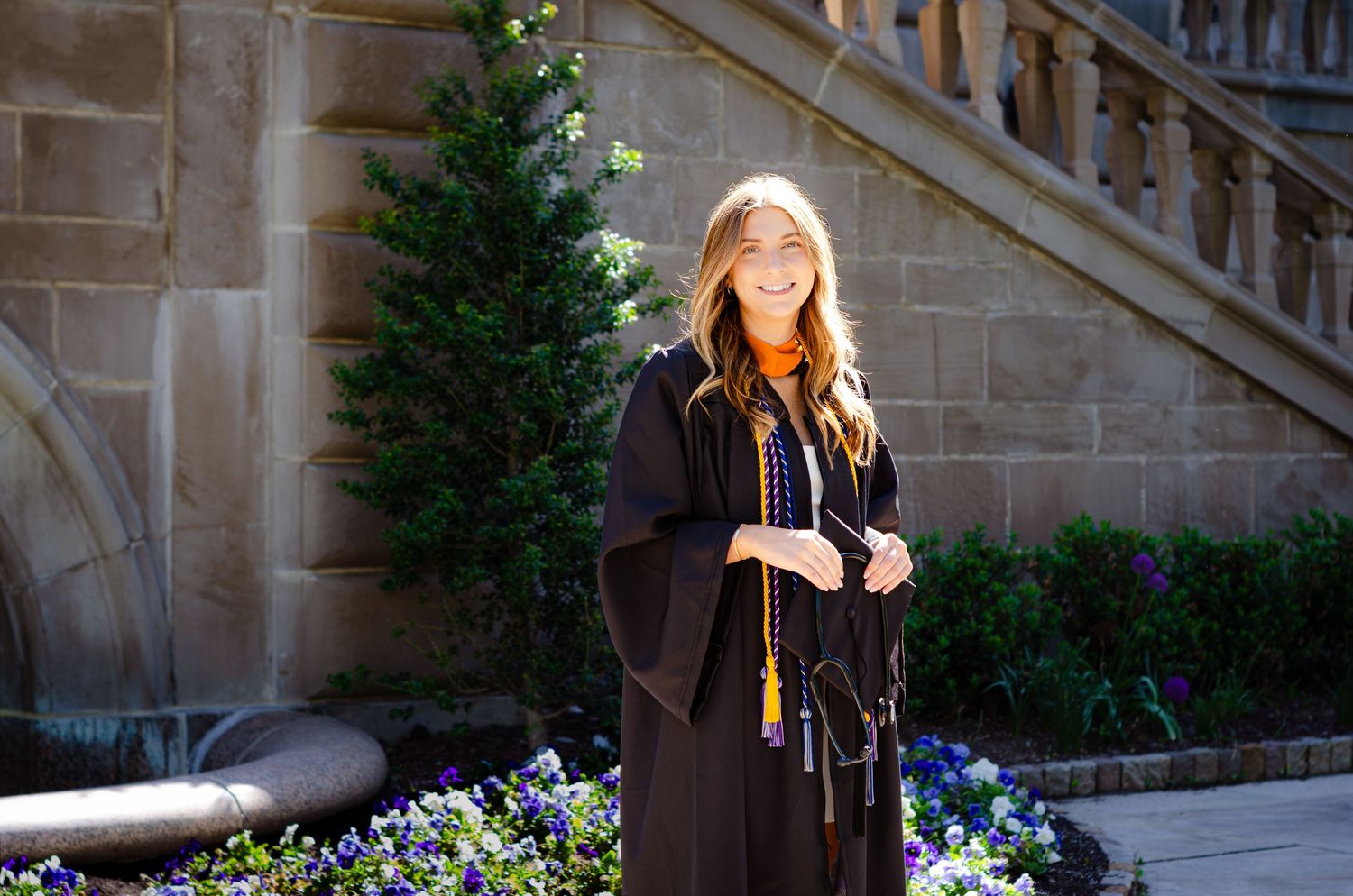 Young woman in black graduation gown with orange sash smiling outdoors near stone building and purple flowers.