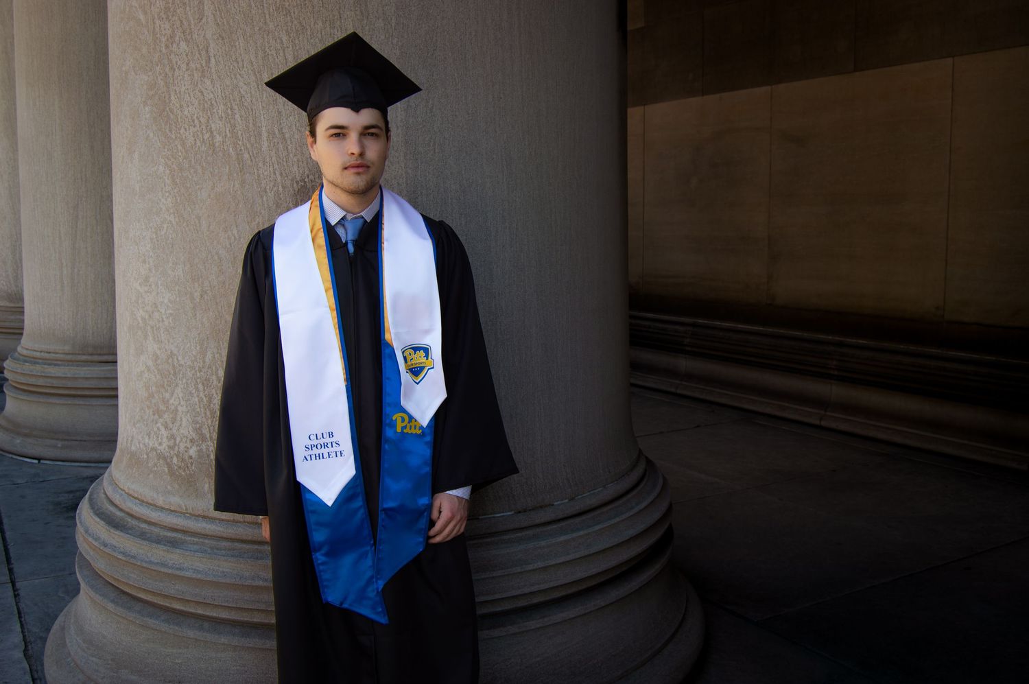 Young man in graduation cap and gown with blue stole poses between stone columns for graduation photo.