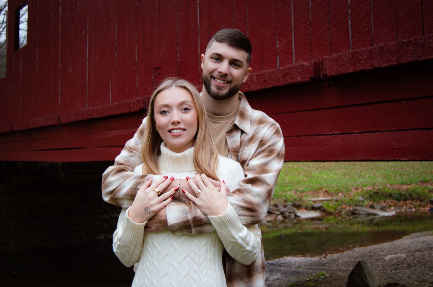 Couple in casual sweaters embraces against red barn wall during outdoor autumn photoshoot.