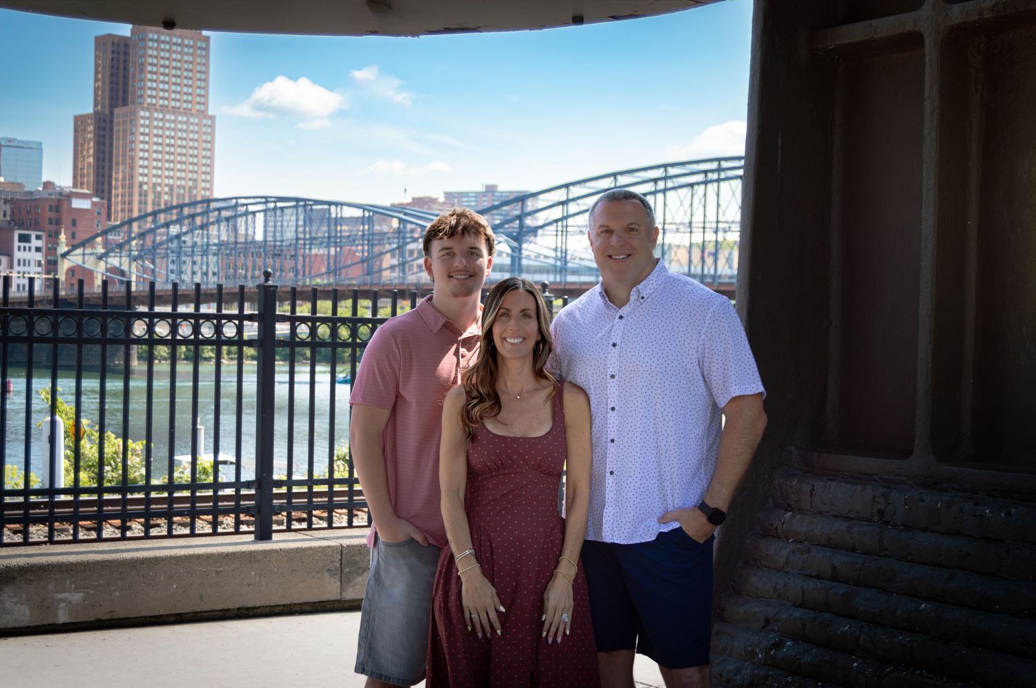 Three people smiling together on outdoor terrace with city skyline and river bridge in background.