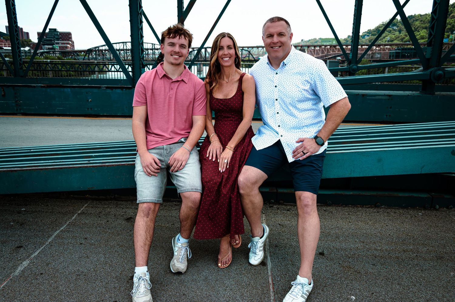 Three smiling people sitting together outdoors on a bridge with a river view behind them.
