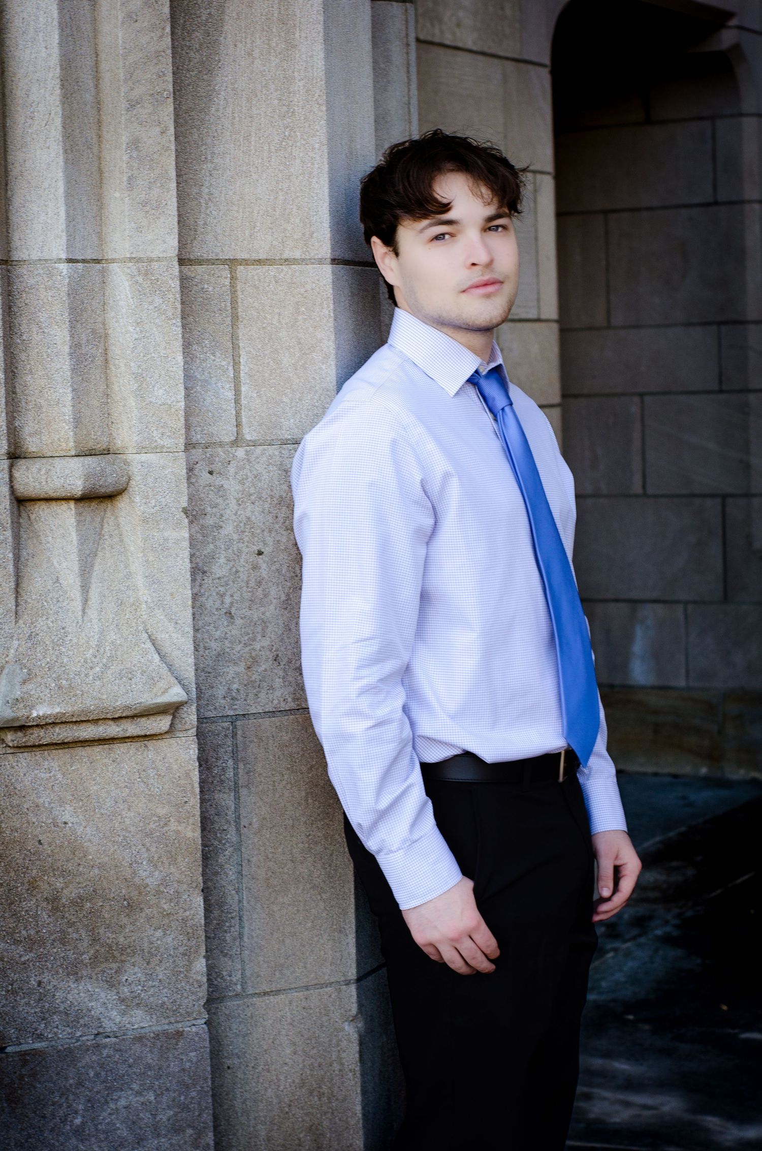 Young man in white dress shirt and blue tie leaning against stone pillar in professional portrait photo.