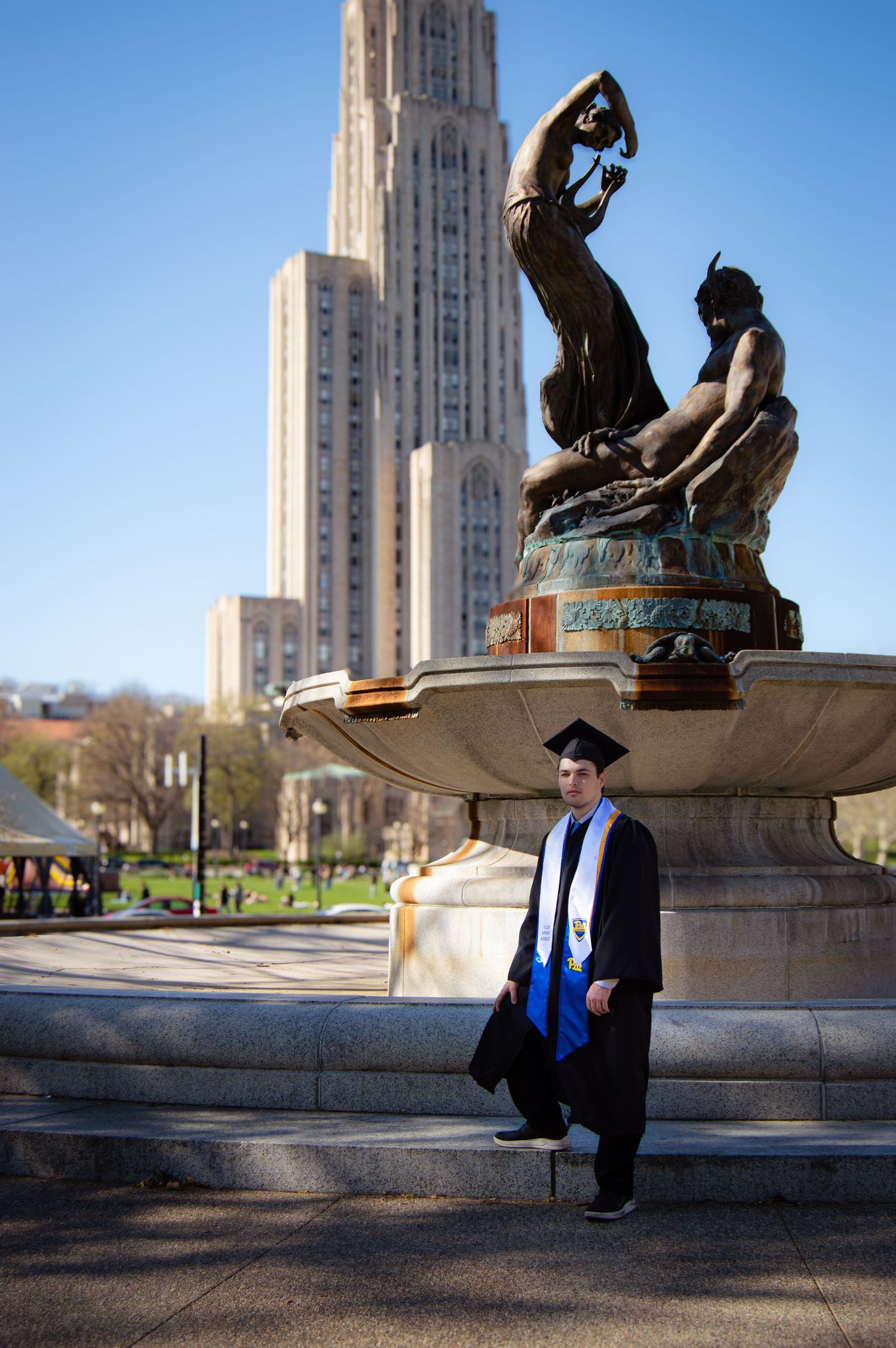 Graduate in cap and gown posing by fountain with Cathedral of Learning in background.
