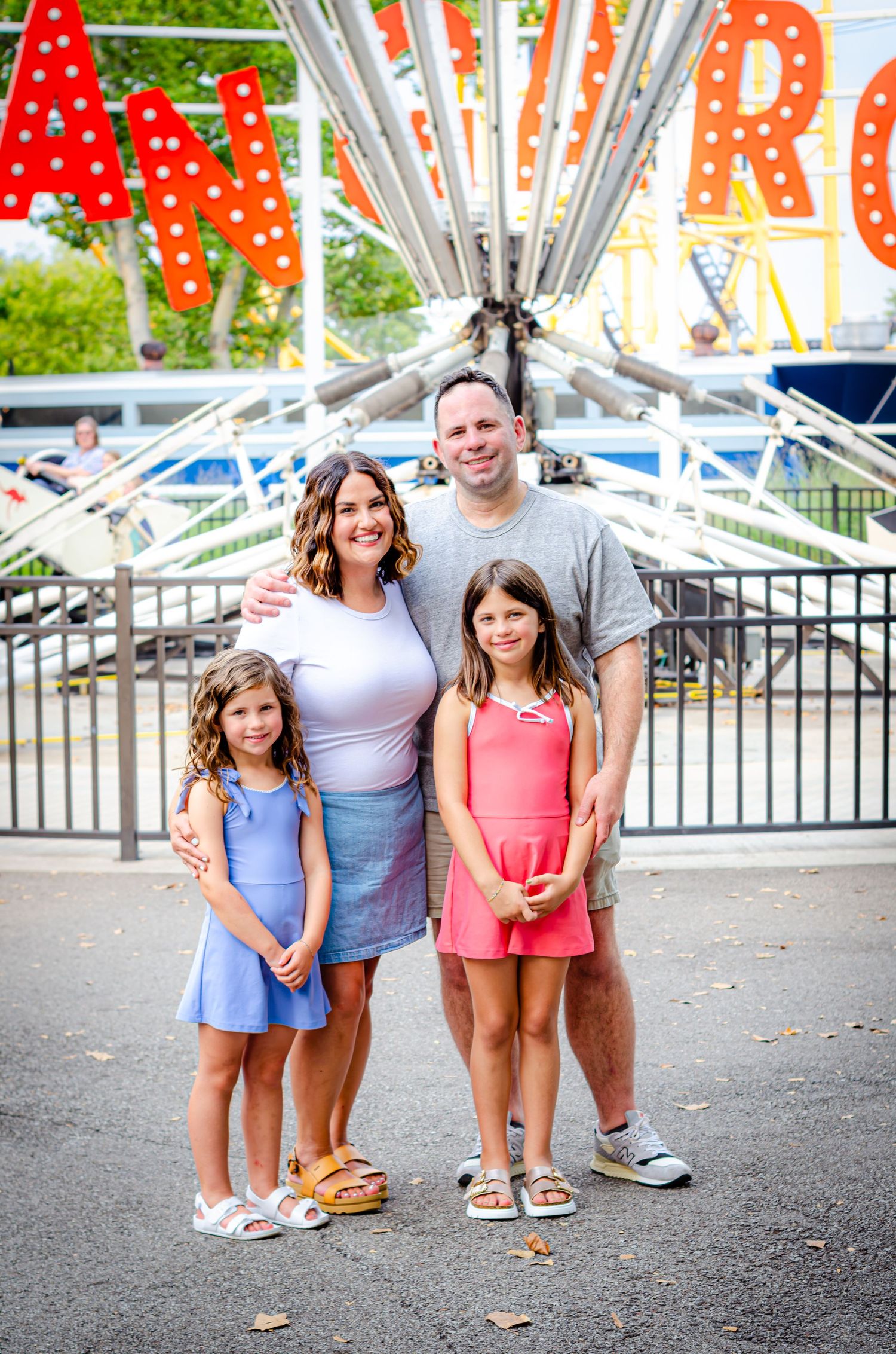 Family of four smiling together at an amusement park, with colorful rides and signs in the background.
