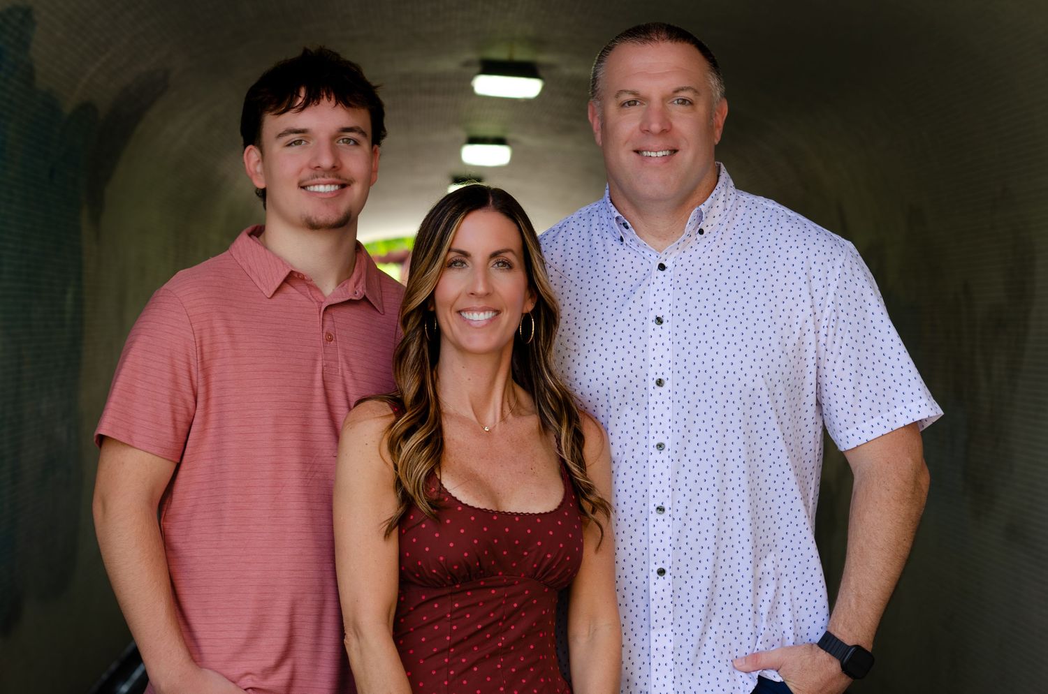 Three smiling people posing together in a tunnel, wearing casual attire in a warm, well-lit setting.