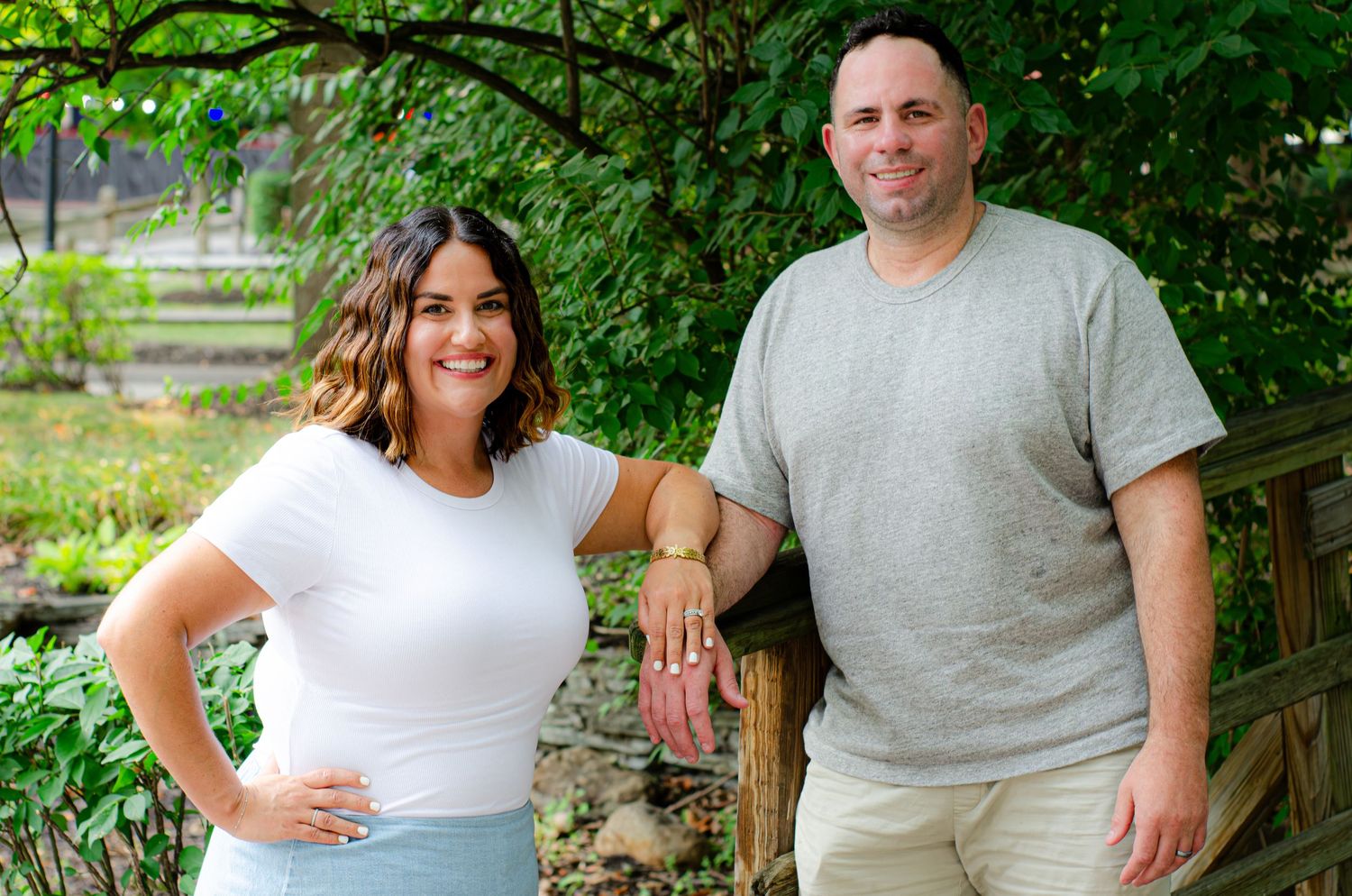 A smiling couple poses outdoors near lush green trees, woman in white top and man in gray shirt.