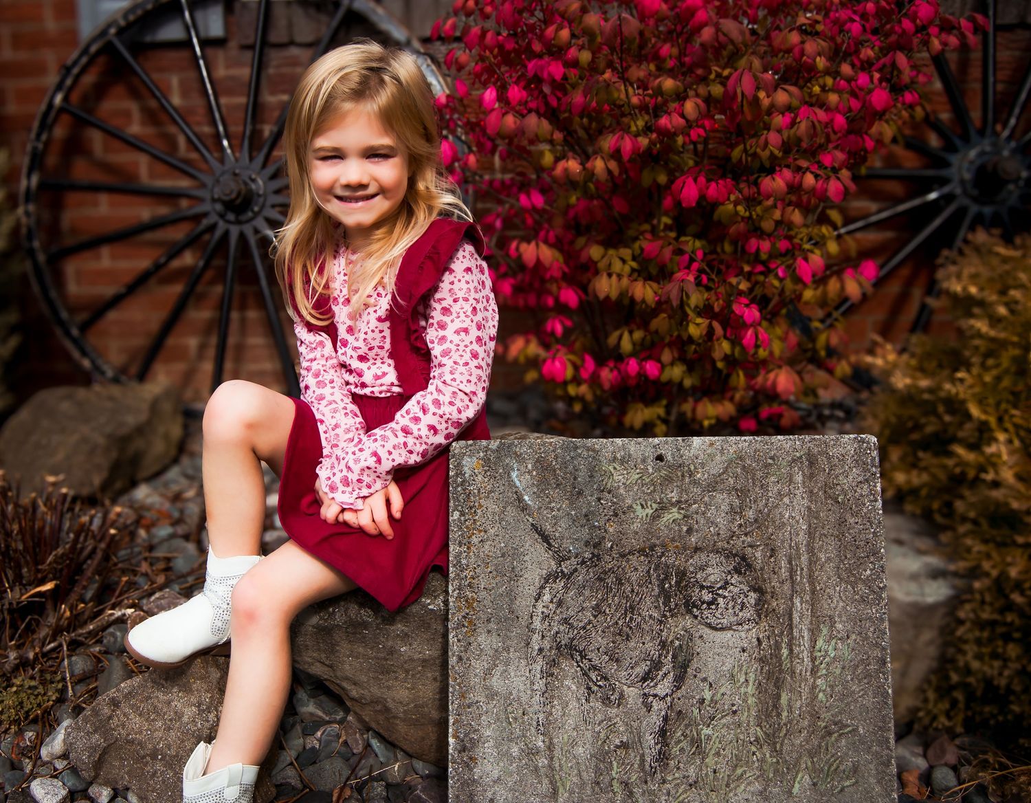 A young child in a burgundy dress sits on a stone wall with red autumn foliage and wagon wheels in the background.