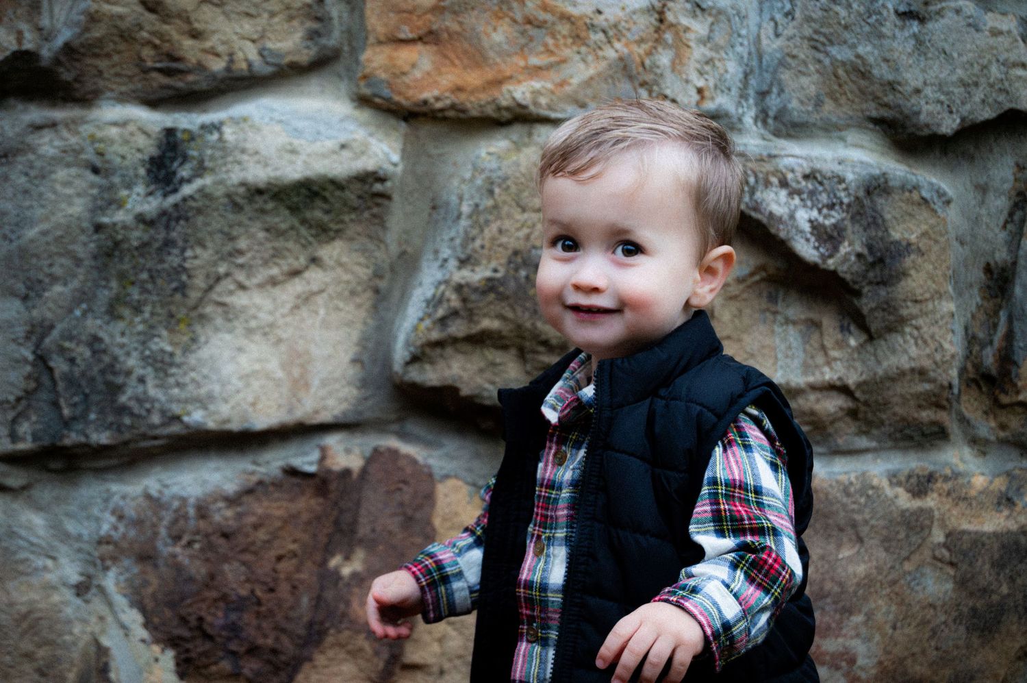 A young child in plaid and black clothing poses playfully against a rustic stone wall background.