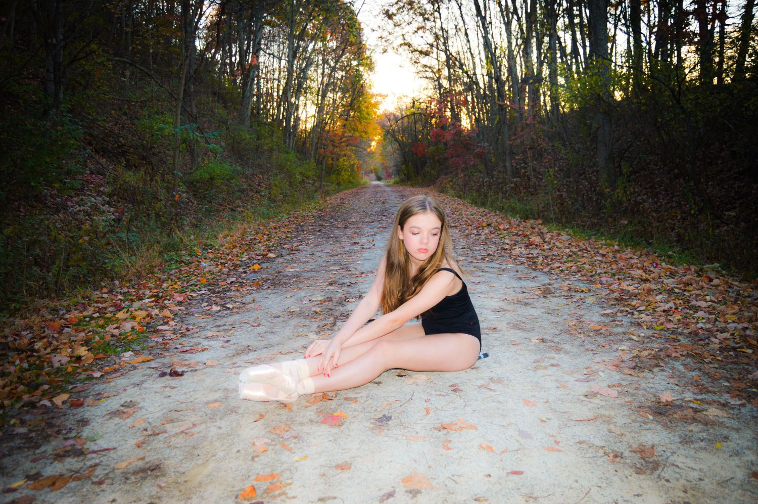 A dancer in ballet attire poses gracefully on a leaf-covered forest path during golden hour sunset.