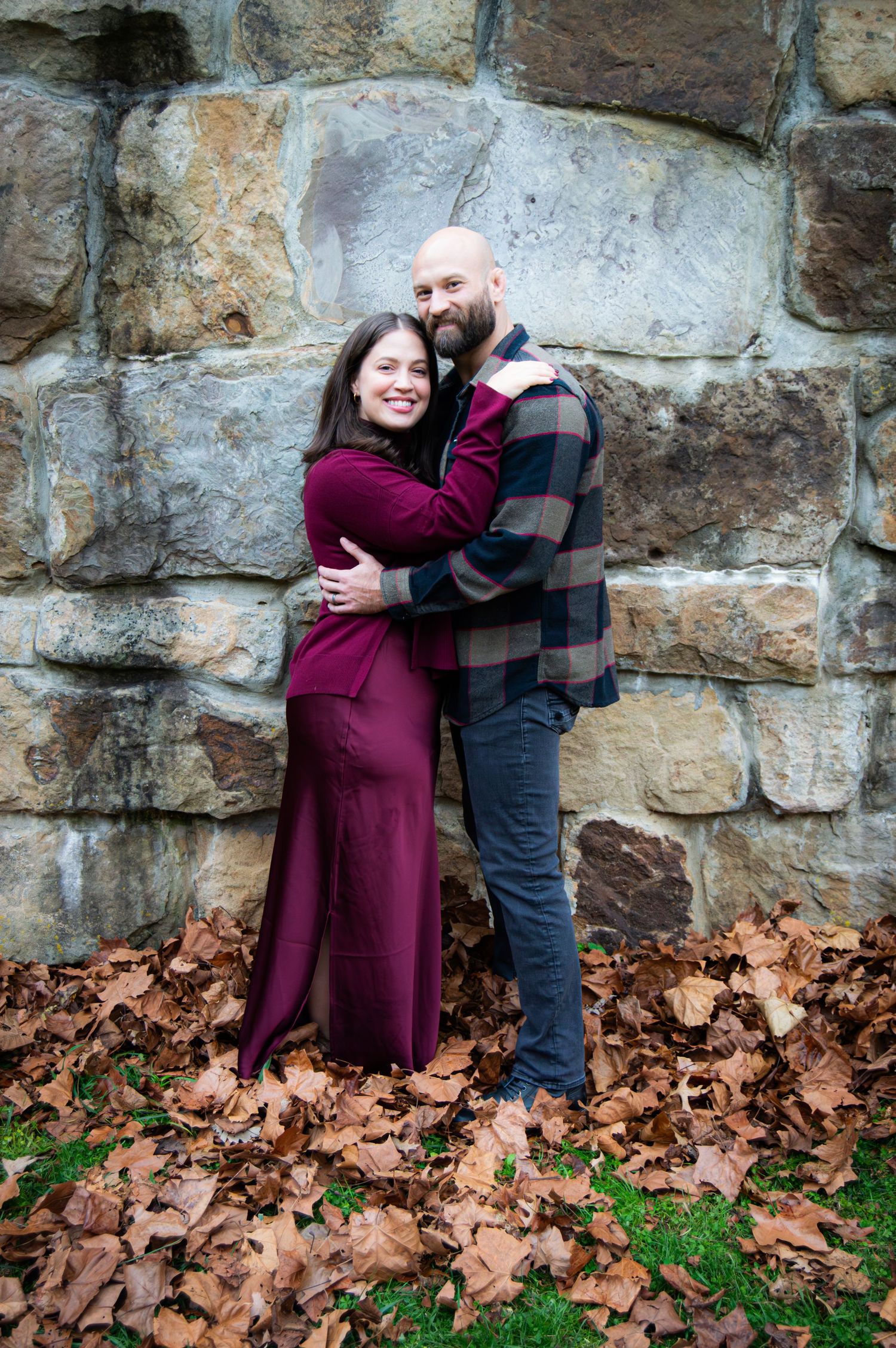A couple shares a tender moment against a stone wall backdrop with autumn leaves scattered on the ground.