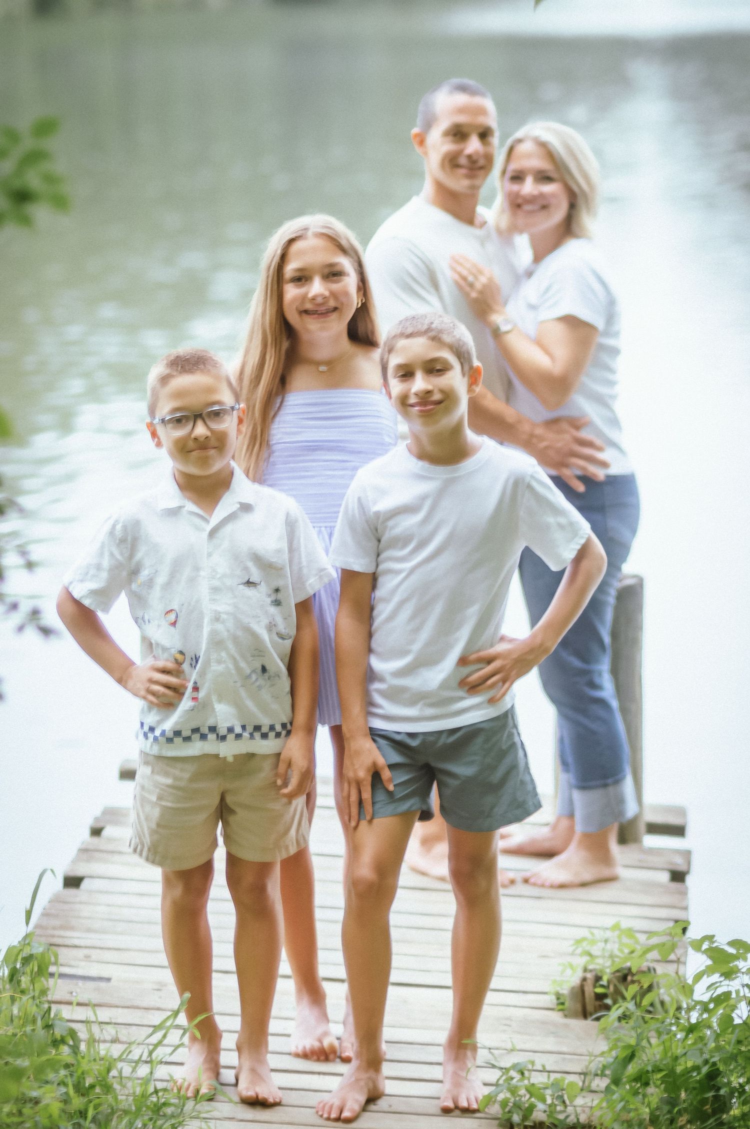 Family group stands together on wooden dock by tranquil lake during outdoor summer photoshoot.