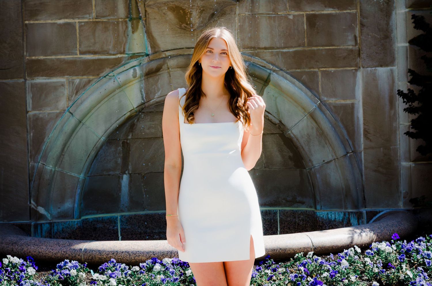Young woman in white mini dress standing before stone fountain surrounded by purple flowers.