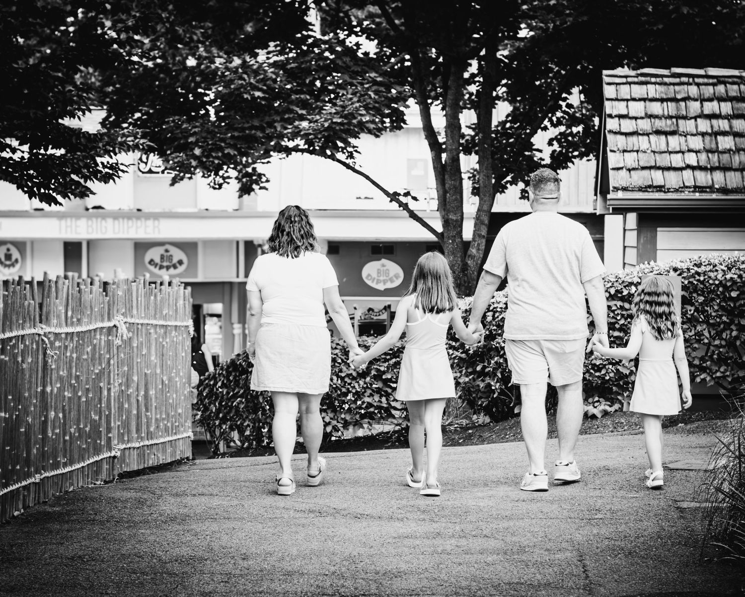 Black and white photo of a family holding hands walking down a path toward a building surrounded by trees.