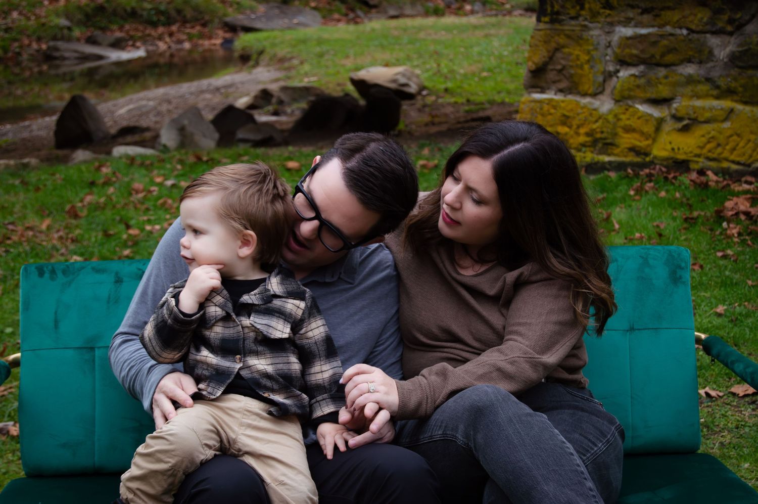 Three people share a loving moment together while sitting on a teal colored bench in an outdoor garden setting.