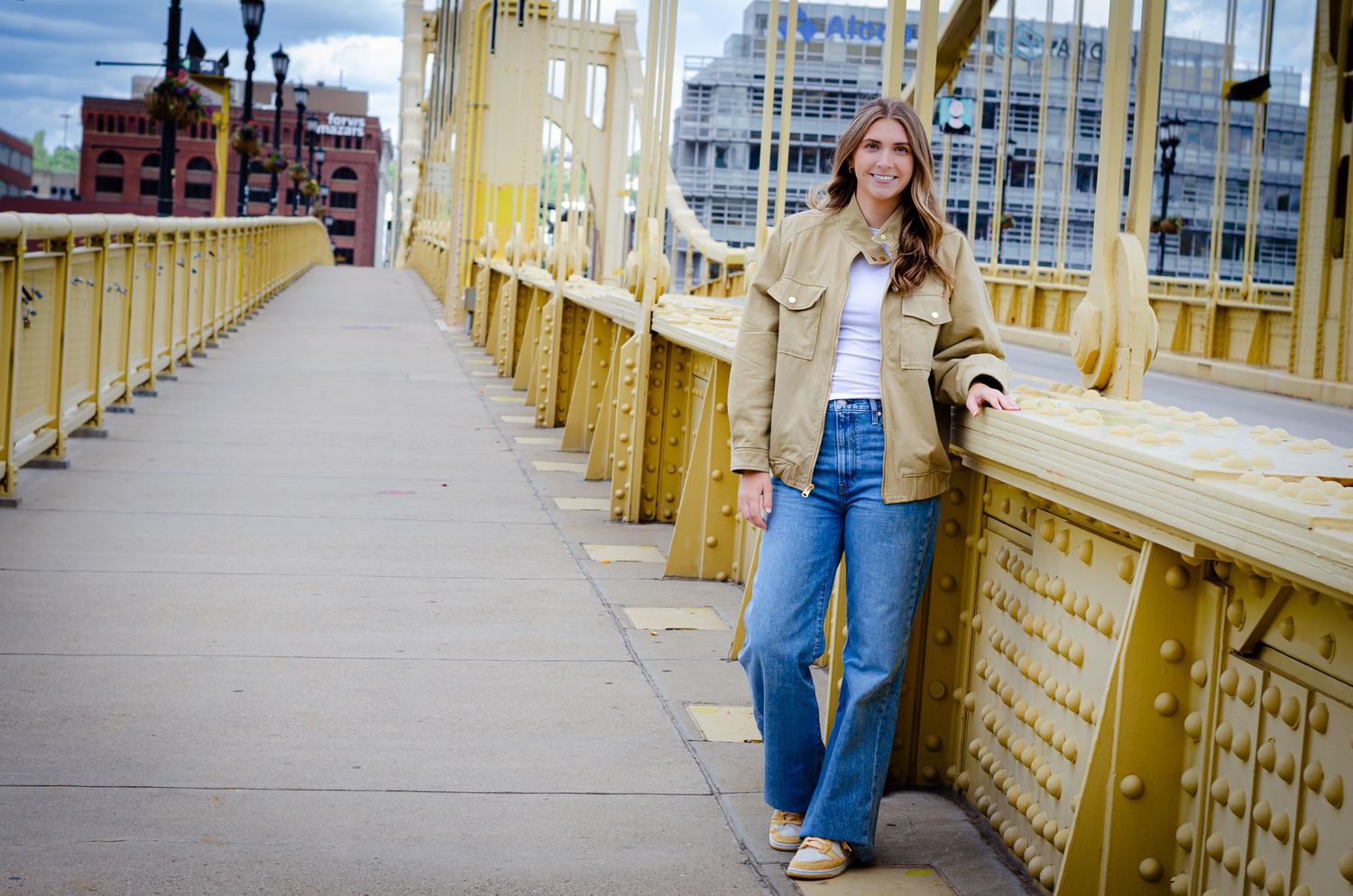 Young woman in tan jacket and wide-leg jeans posing on yellow steel bridge in urban setting.