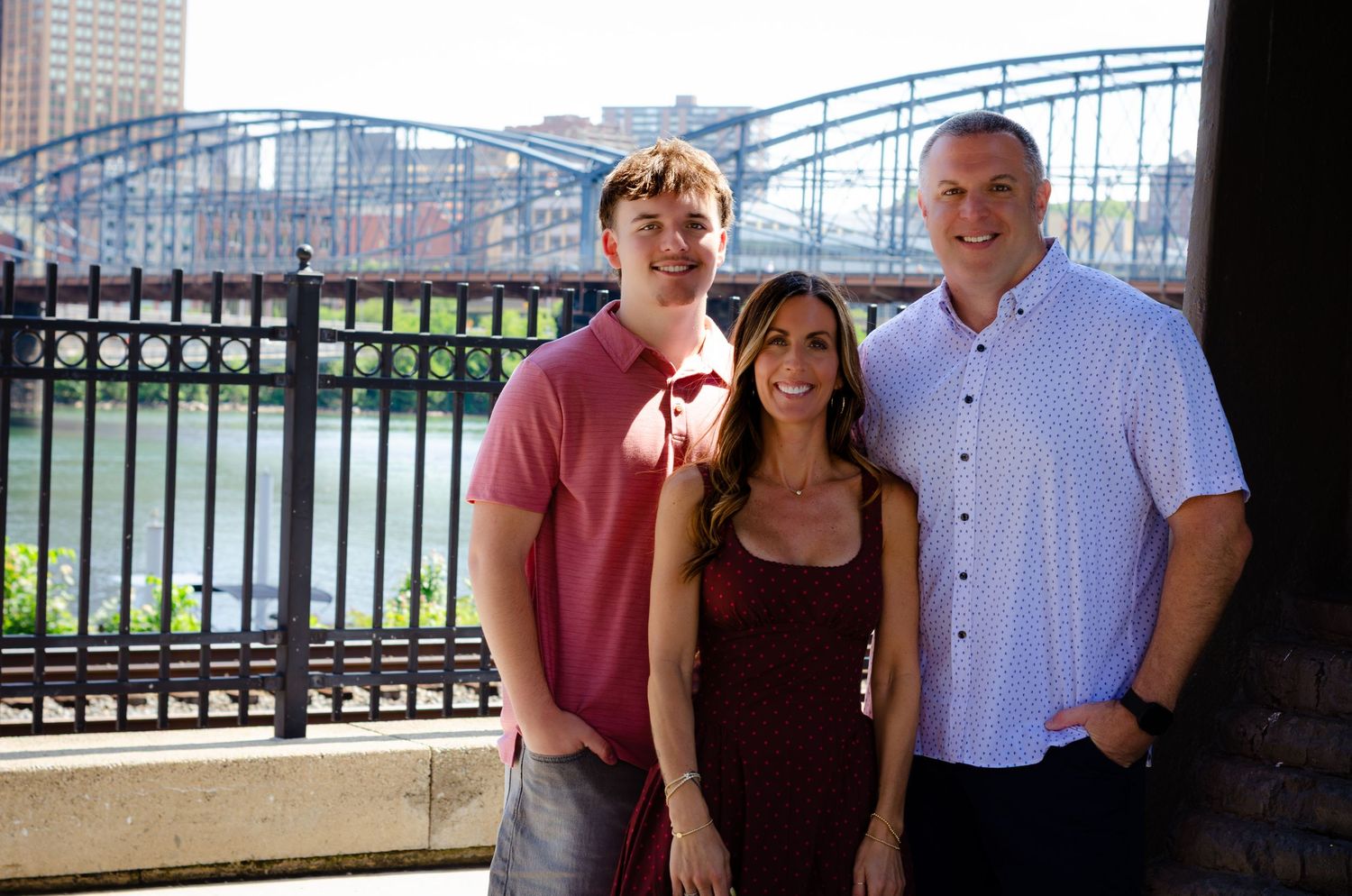 Three smiling people posing outdoors near a river with a bridge in the background on a sunny day.