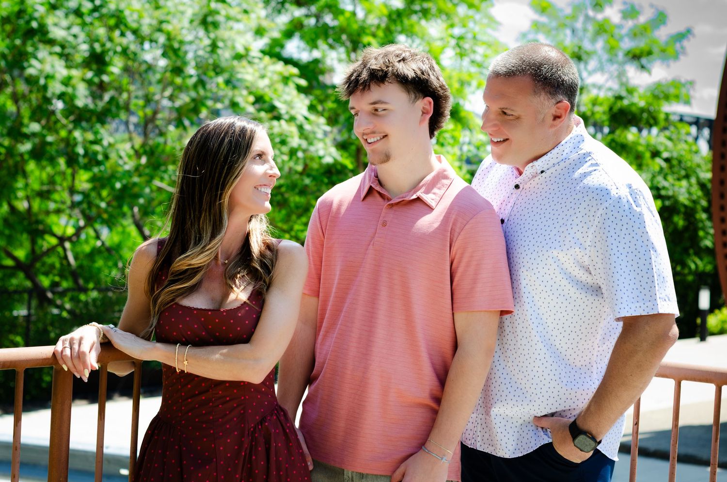 Three smiling people standing together on a wooden deck surrounded by lush green trees on a sunny day.