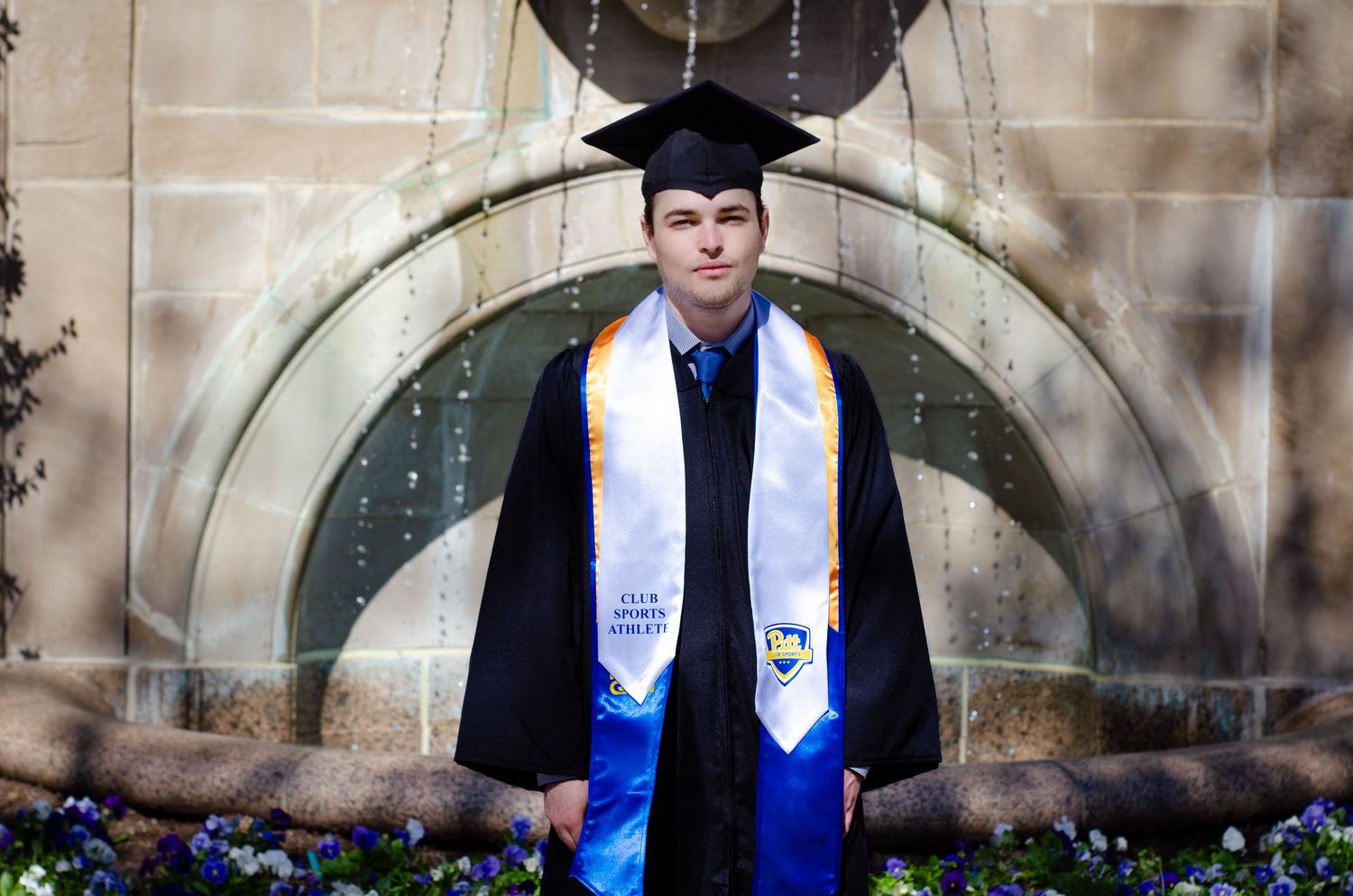 Graduate in black cap and gown with blue and gold stole poses proudly in front of an arched stone building.